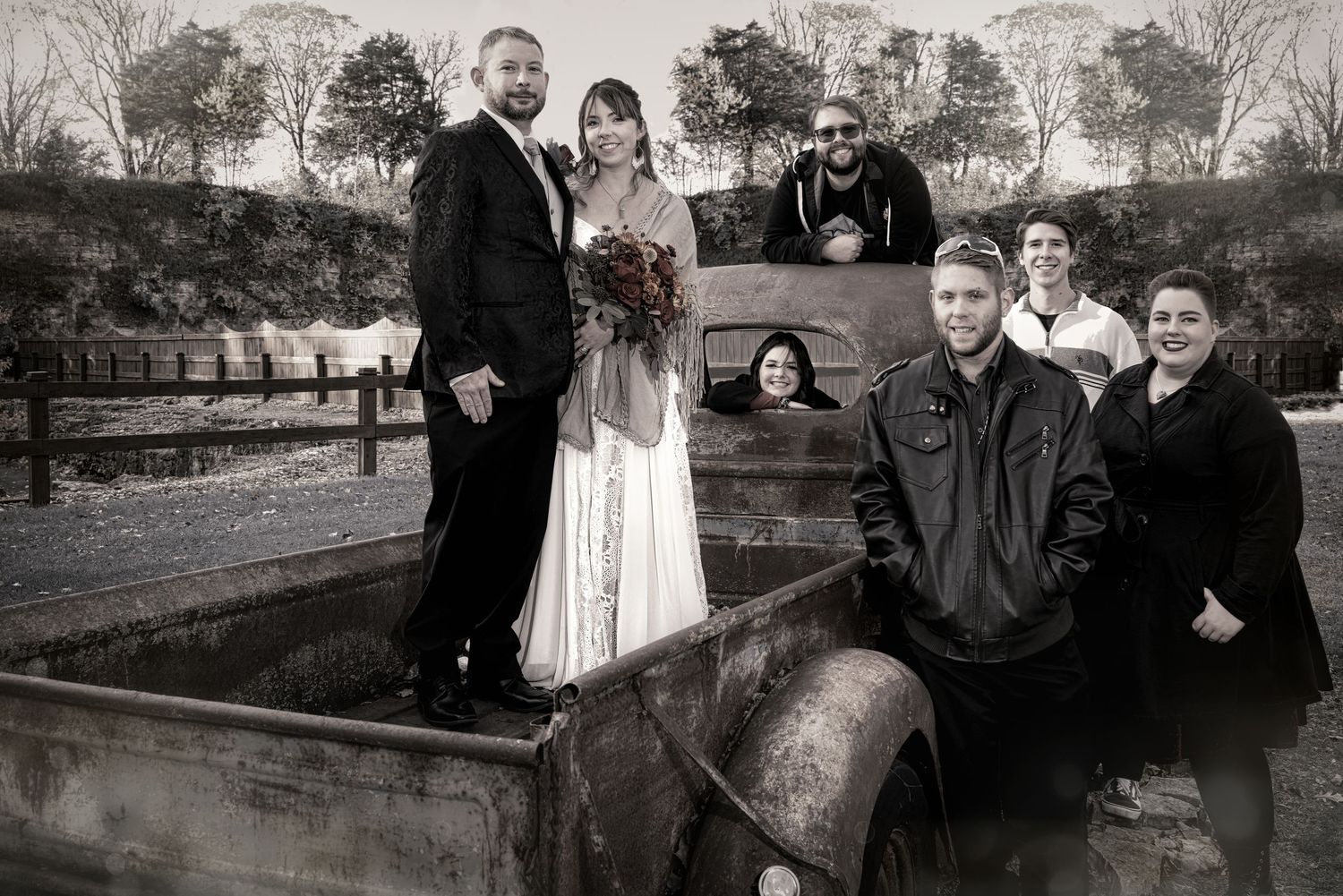 Black and white wedding photo of bride and groom with guests posed around a vintage truck outdoors.