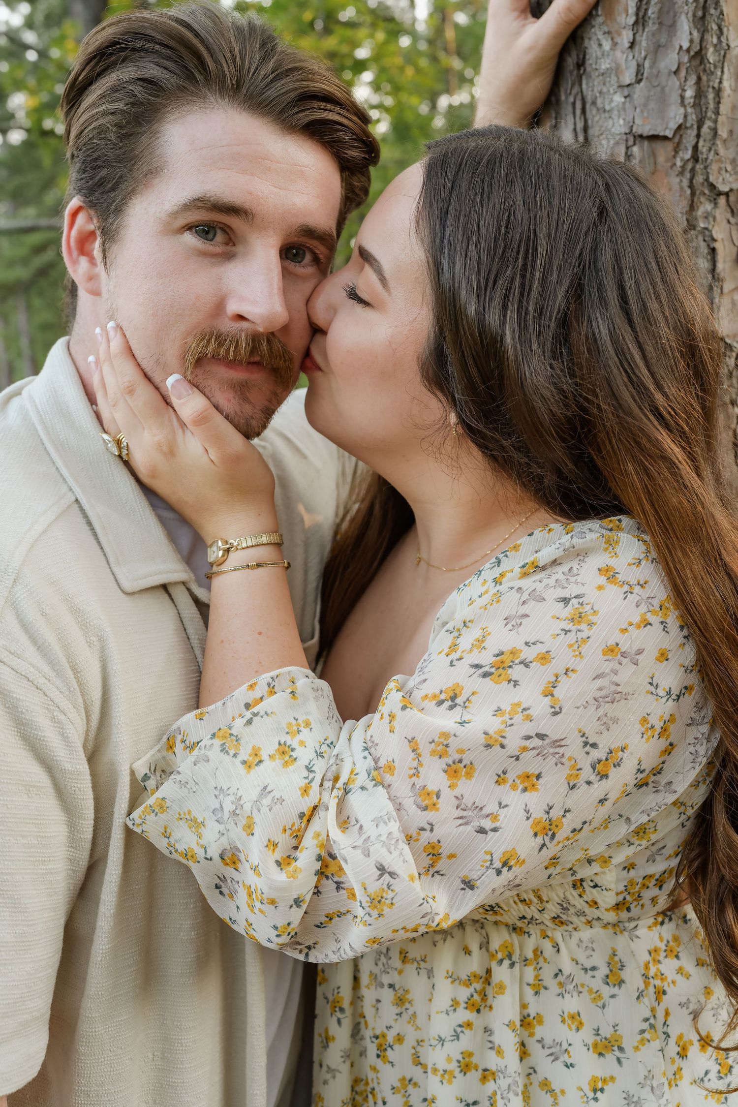 Young woman in floral dress kissing mans cheek as he leans against a tree outdoors.