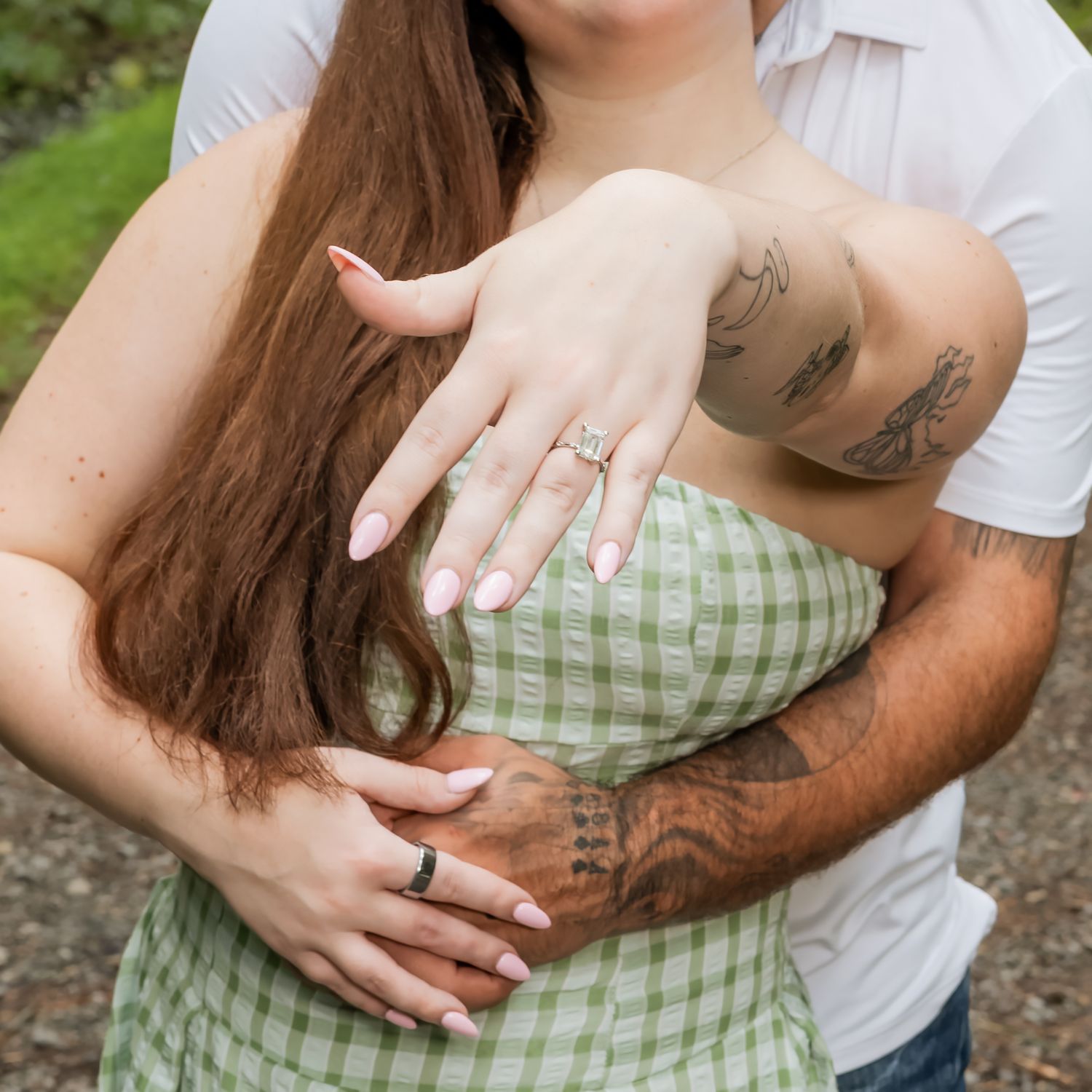 Couple embracing outdoors, woman showing engagement ring, wearing green gingham dress, man with tattoos, romantic proposal moment.