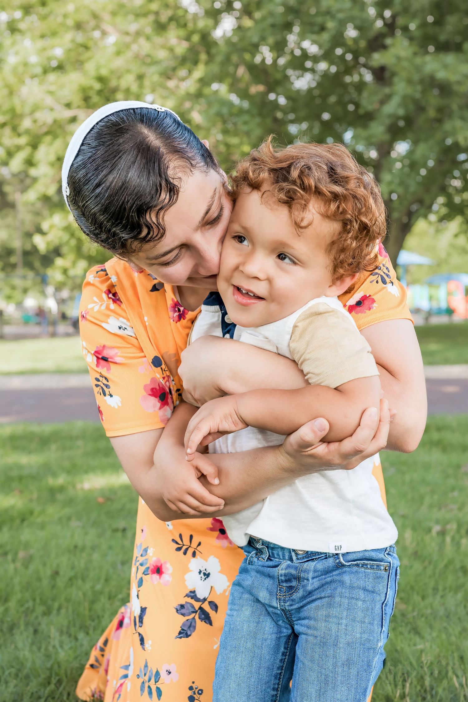 Loving mother in floral dress hugging and kissing curly haired toddler in park.