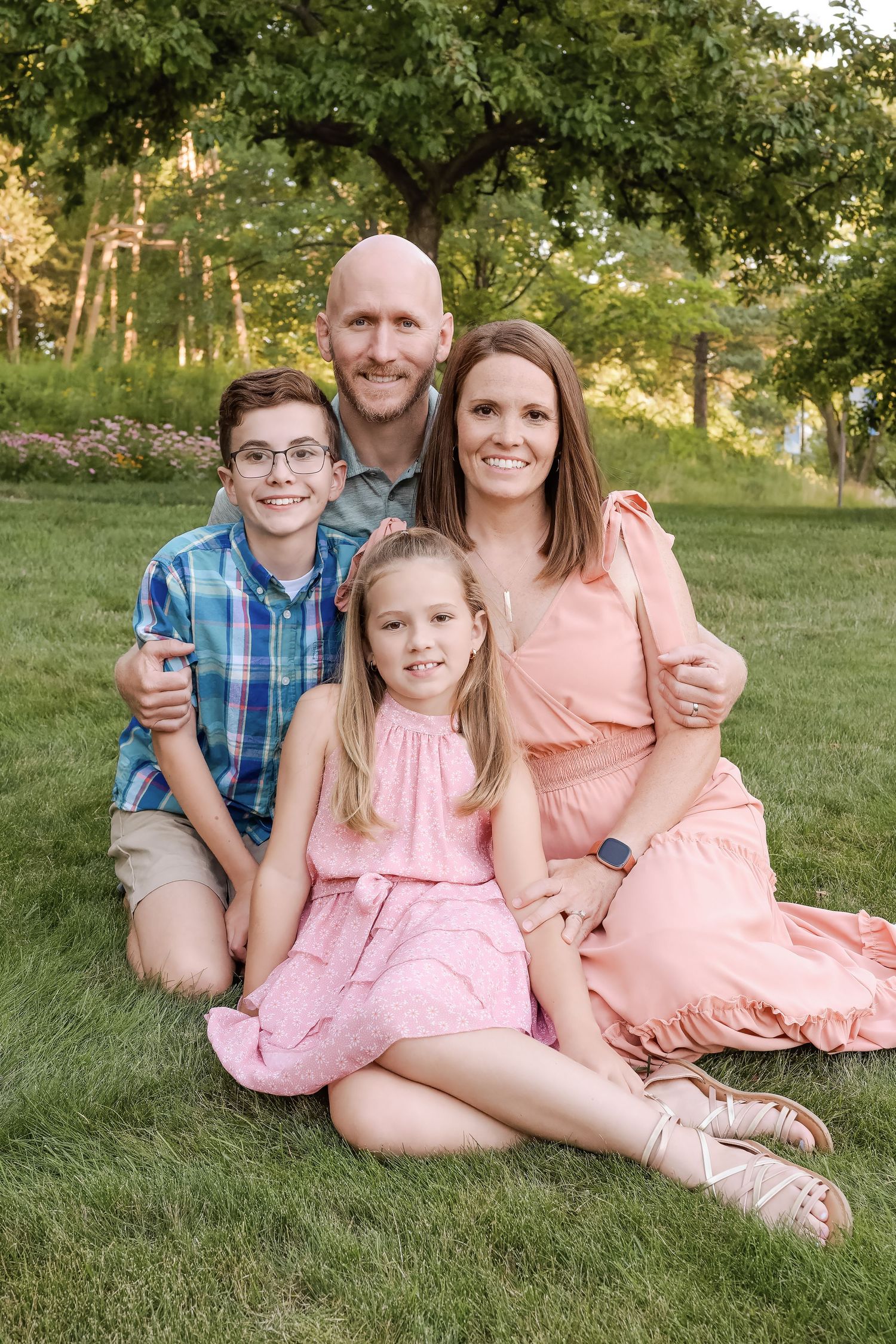 Happy family of four posing on green grass outdoors, parents with son in plaid shirt and daughter in pink dress.