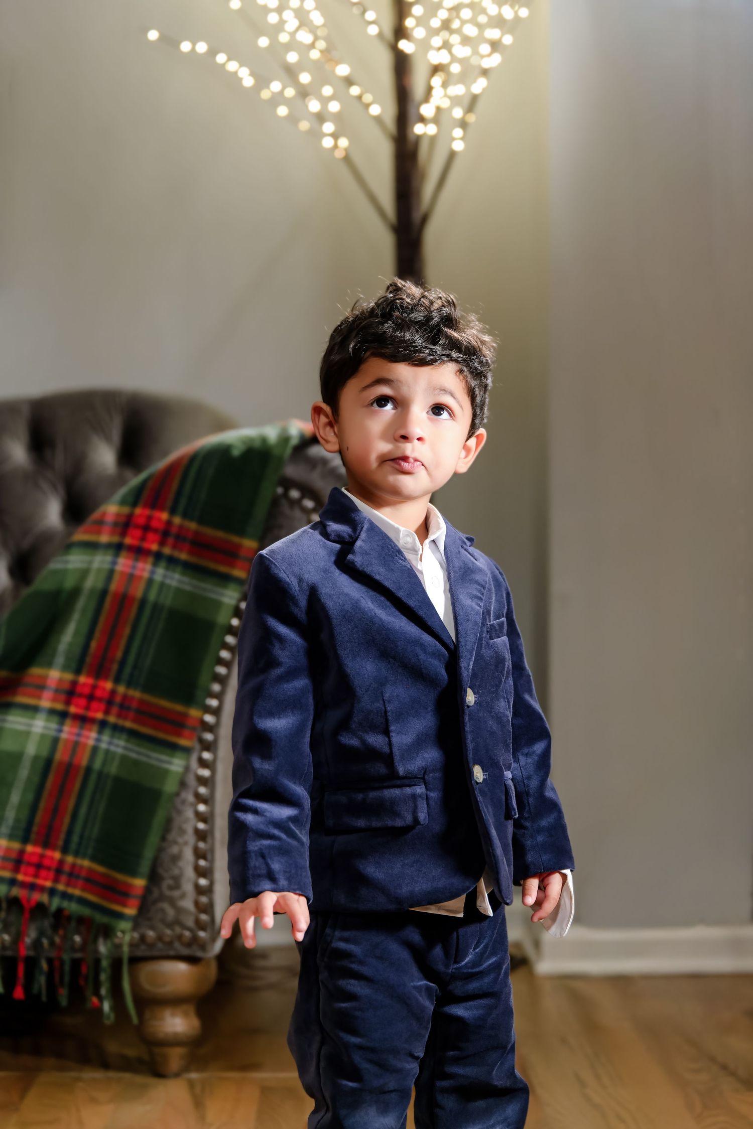 Stylish toddler boy in navy blue suit standing indoors with festive bokeh lights in background.