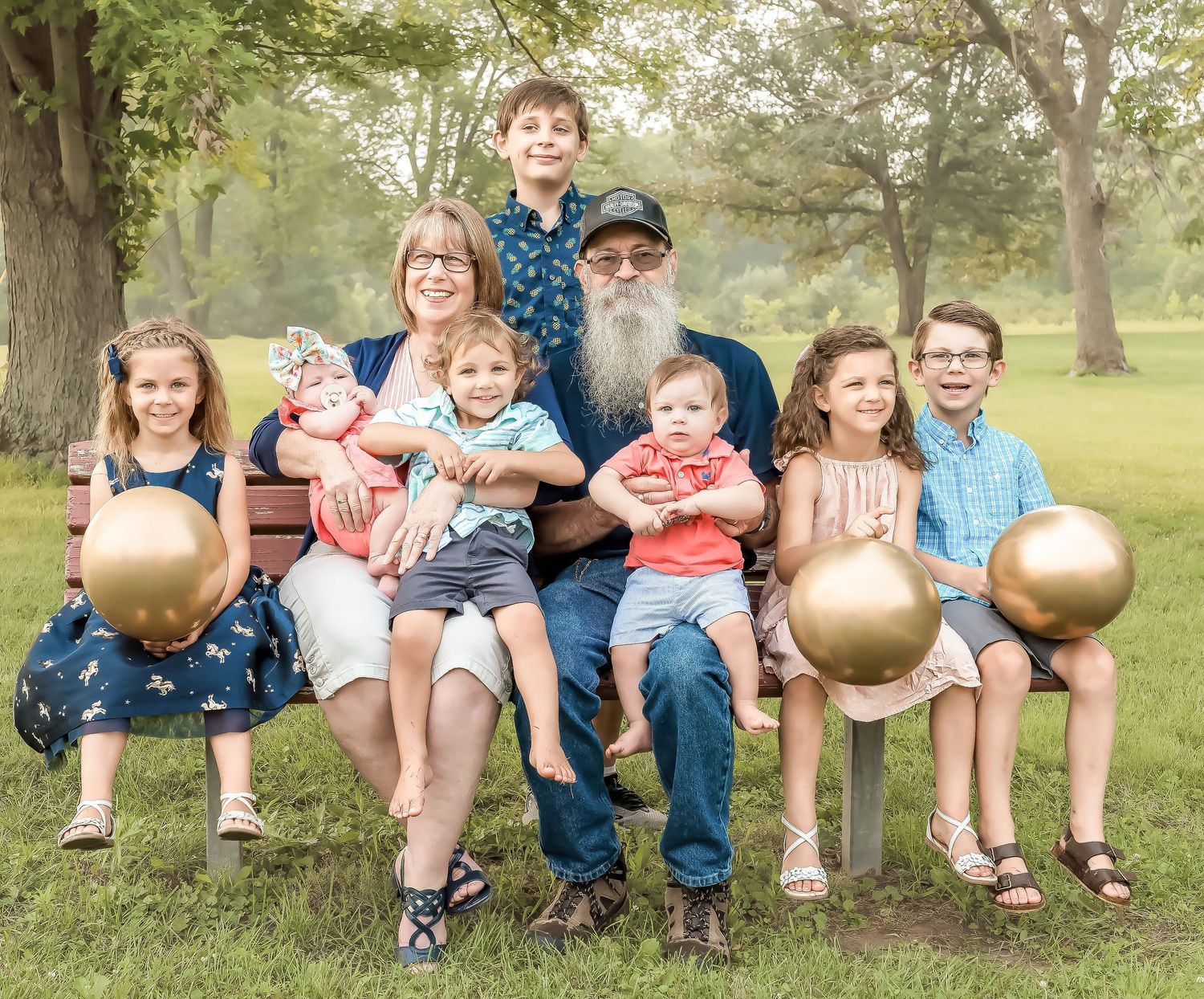 Family portrait of grandparents with seven grandchildren sitting on a bench outdoors in a lush green park.