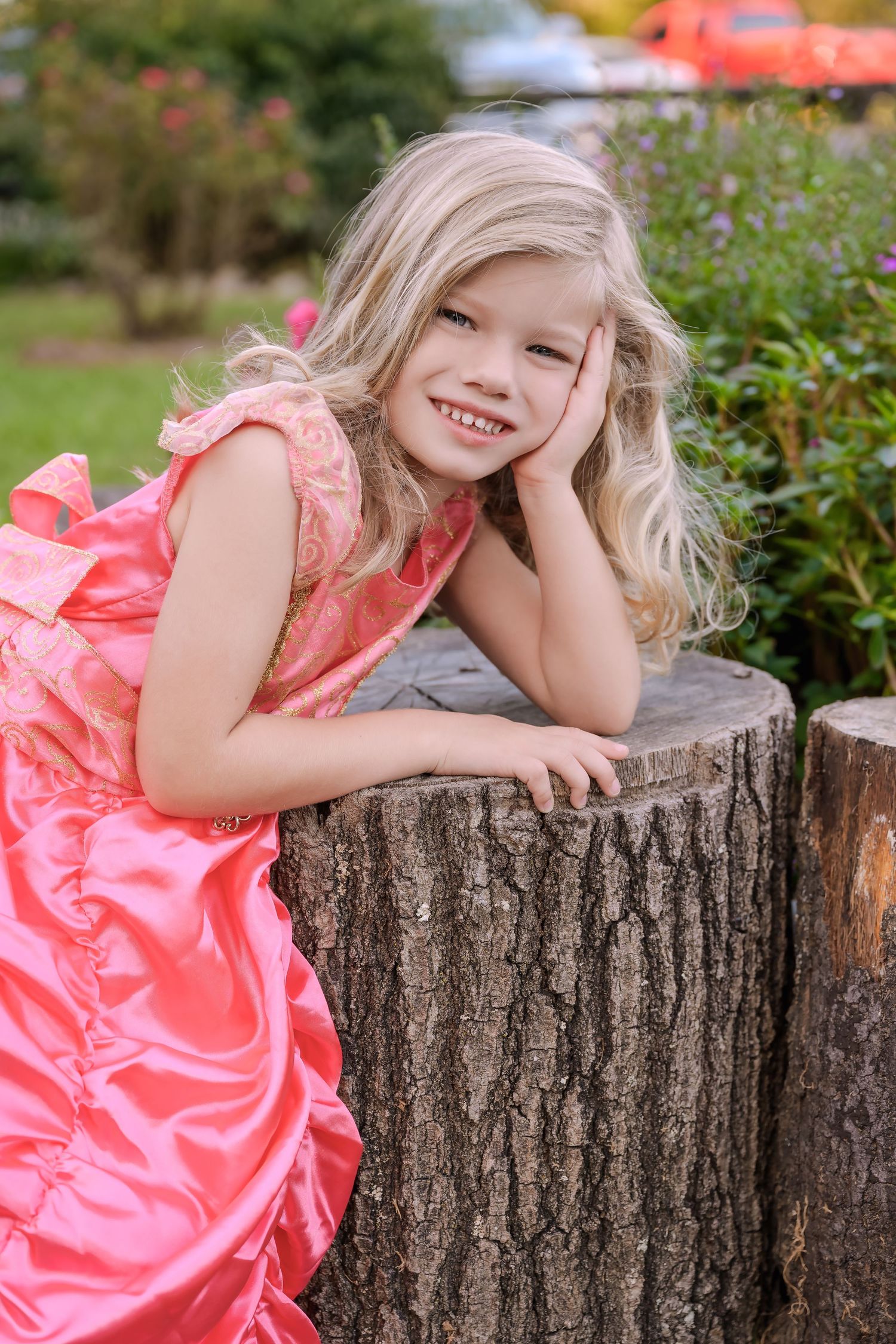 Smiling blonde girl in pink satin dress leaning on rustic tree stump in garden setting.