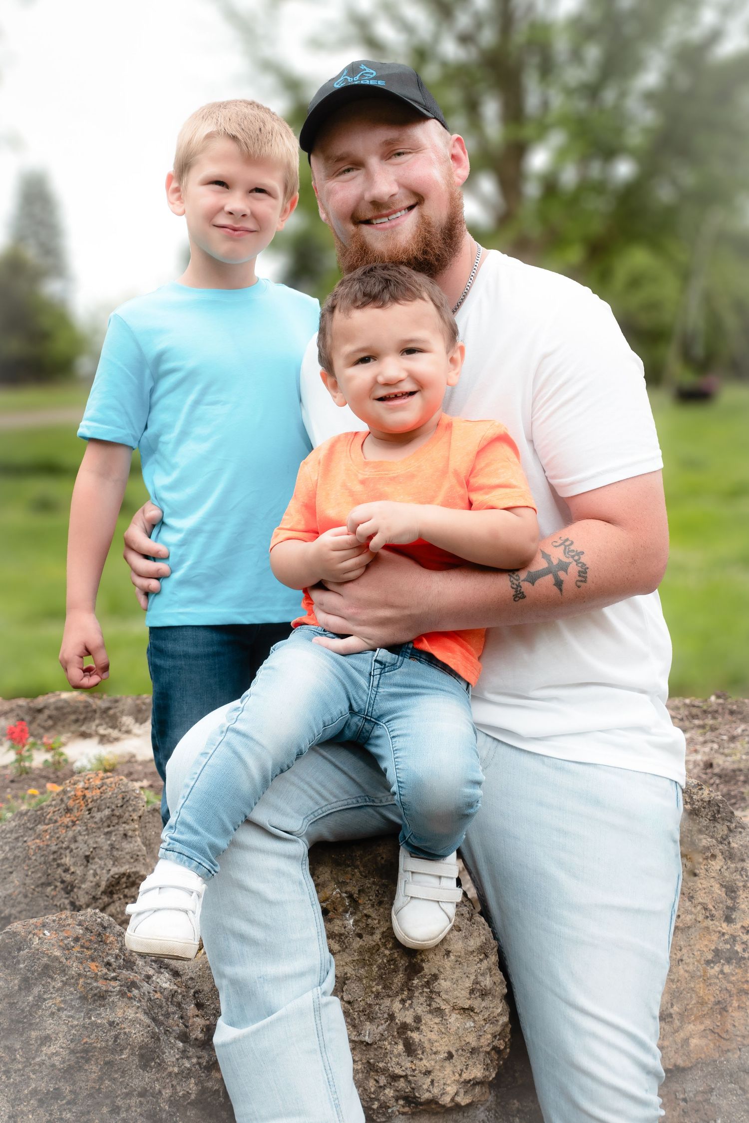 A smiling bearded father in a white tee and cap poses outdoors with his two young sons in a lush green park.
