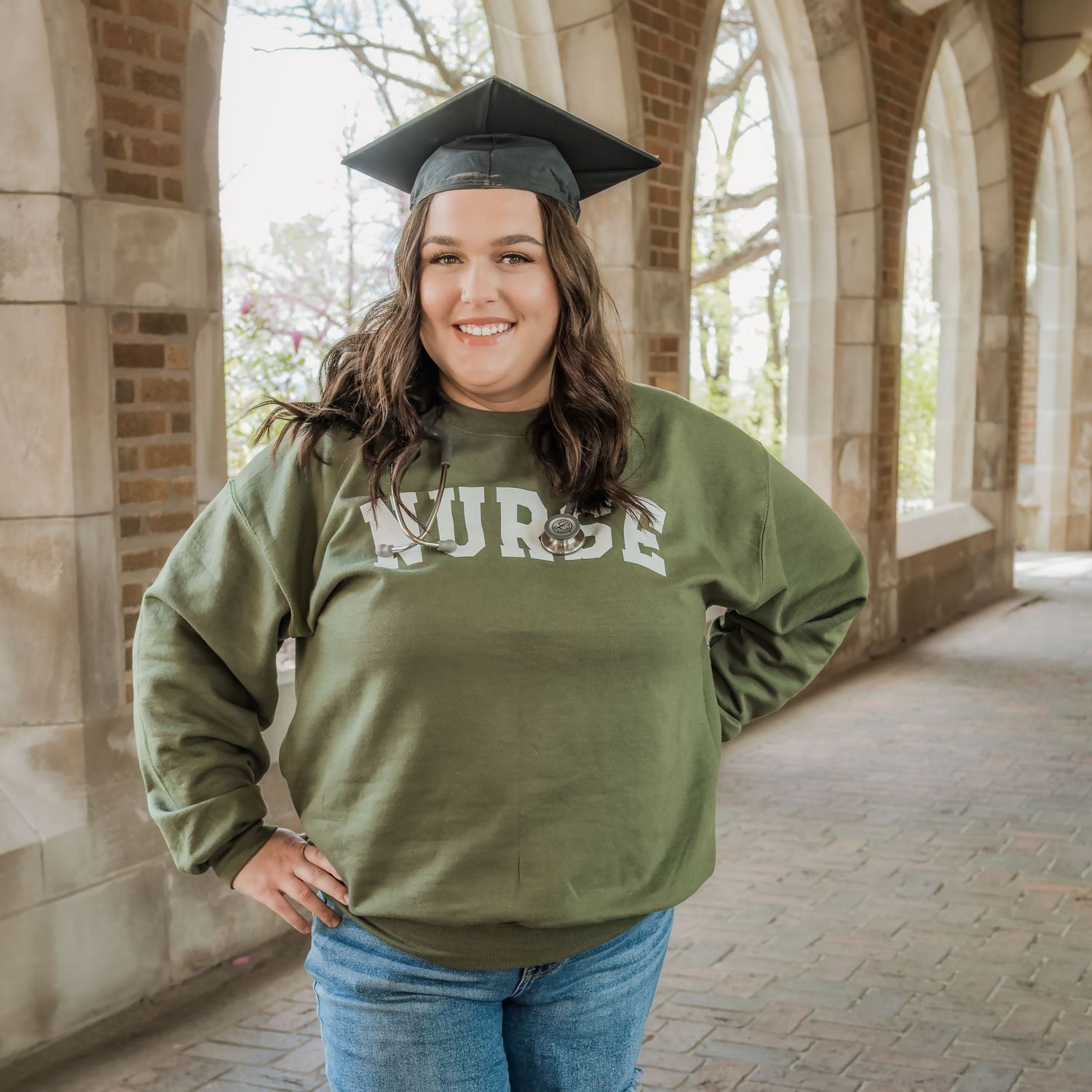 Happy nursing graduate wearing cap and olive green nurse sweatshirt under stone archway.