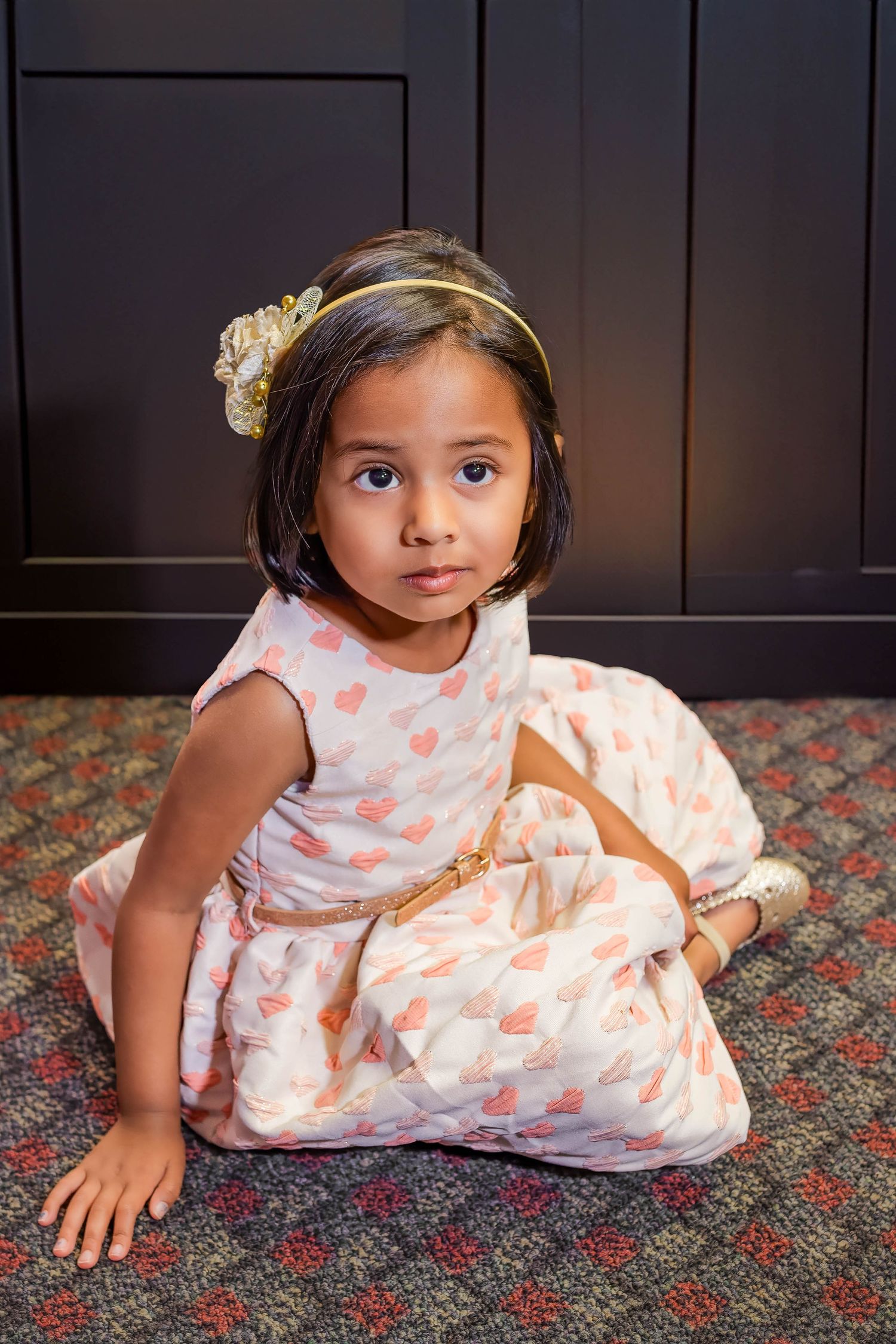 Adorable young girl in white heart-print dress with gold headband sitting on patterned carpet floor.
