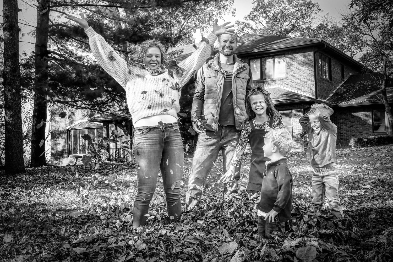 Black and white photo of a joyful family of four playing in fallen leaves outside a rustic home.