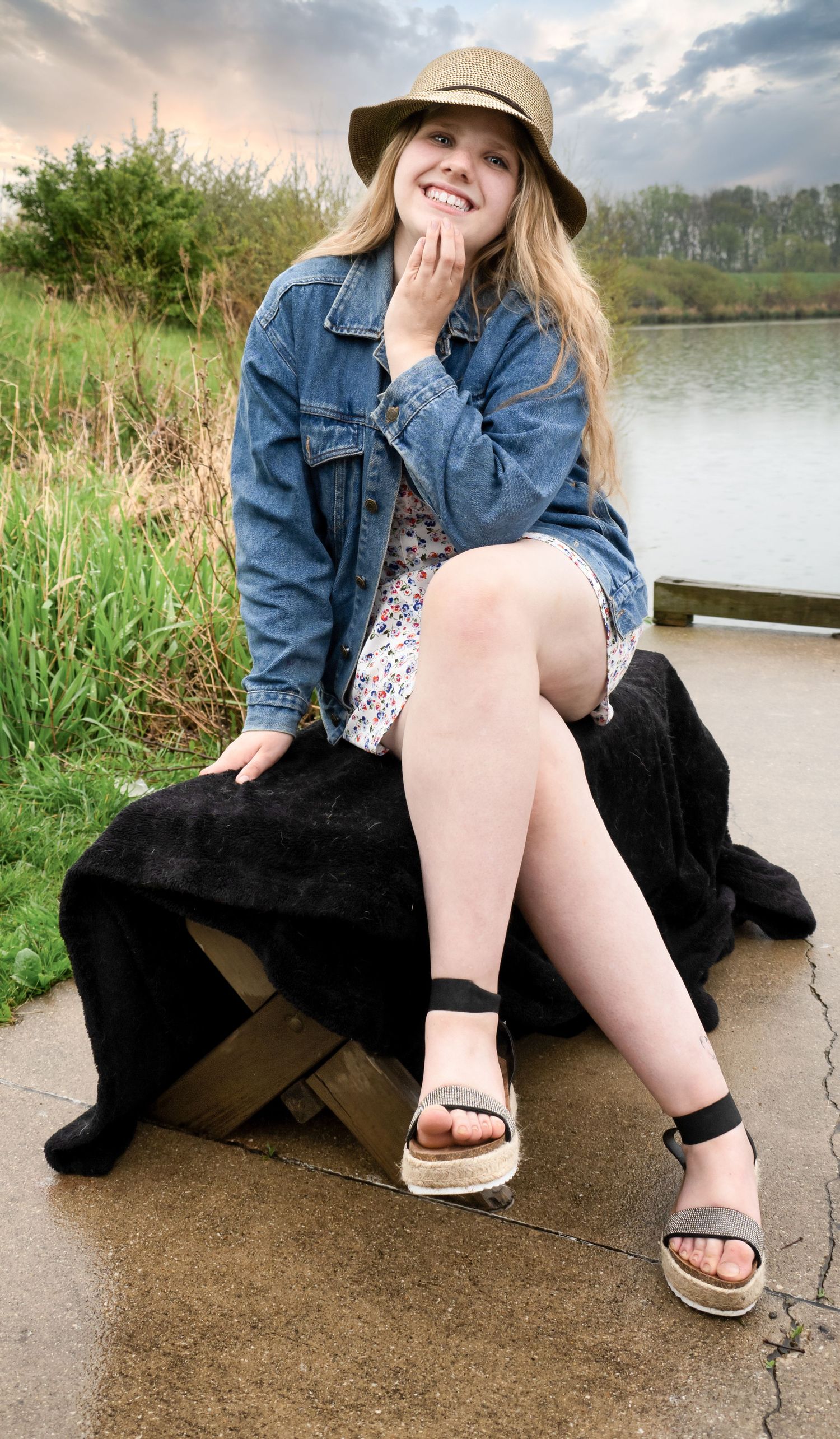 Smiling blonde woman in denim jacket and floral dress wearing hat seated by lake.