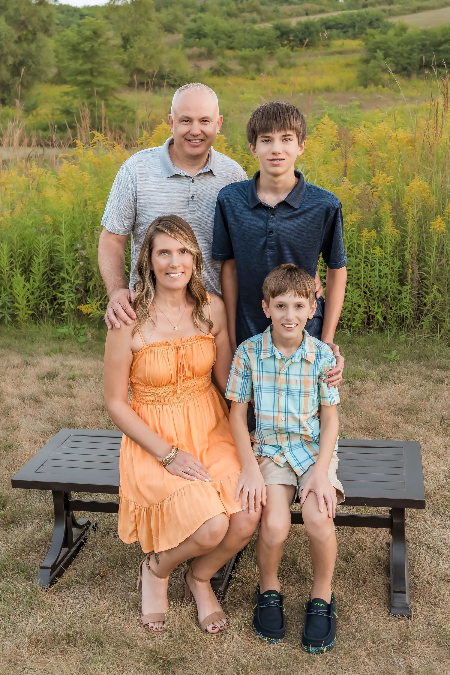 Family of four poses outdoors on a bench surrounded by lush greenery during golden hour.
