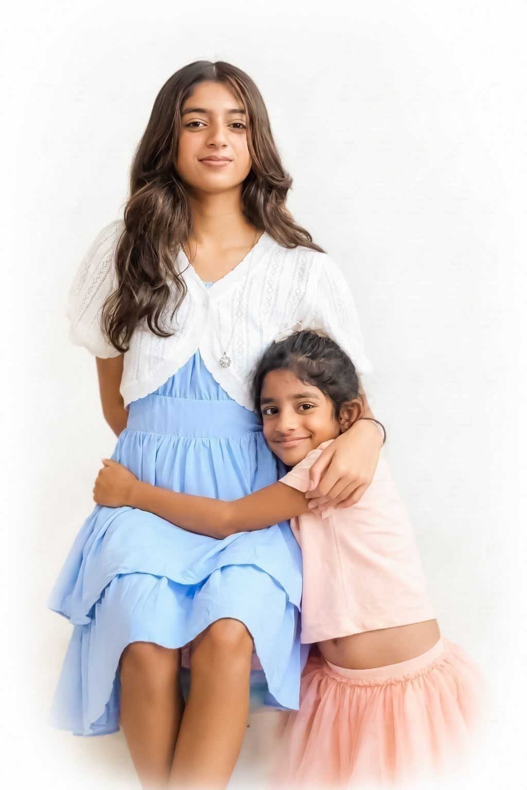 Two young sisters posing together, older girl in blue skirt and white top, younger in pink outfit, studio portrait.
