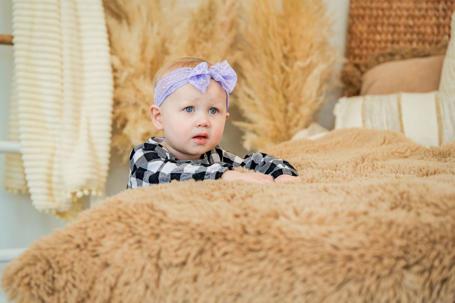 Adorable baby girl wearing a purple bow headband and plaid outfit, peeking over a fluffy tan blanket.