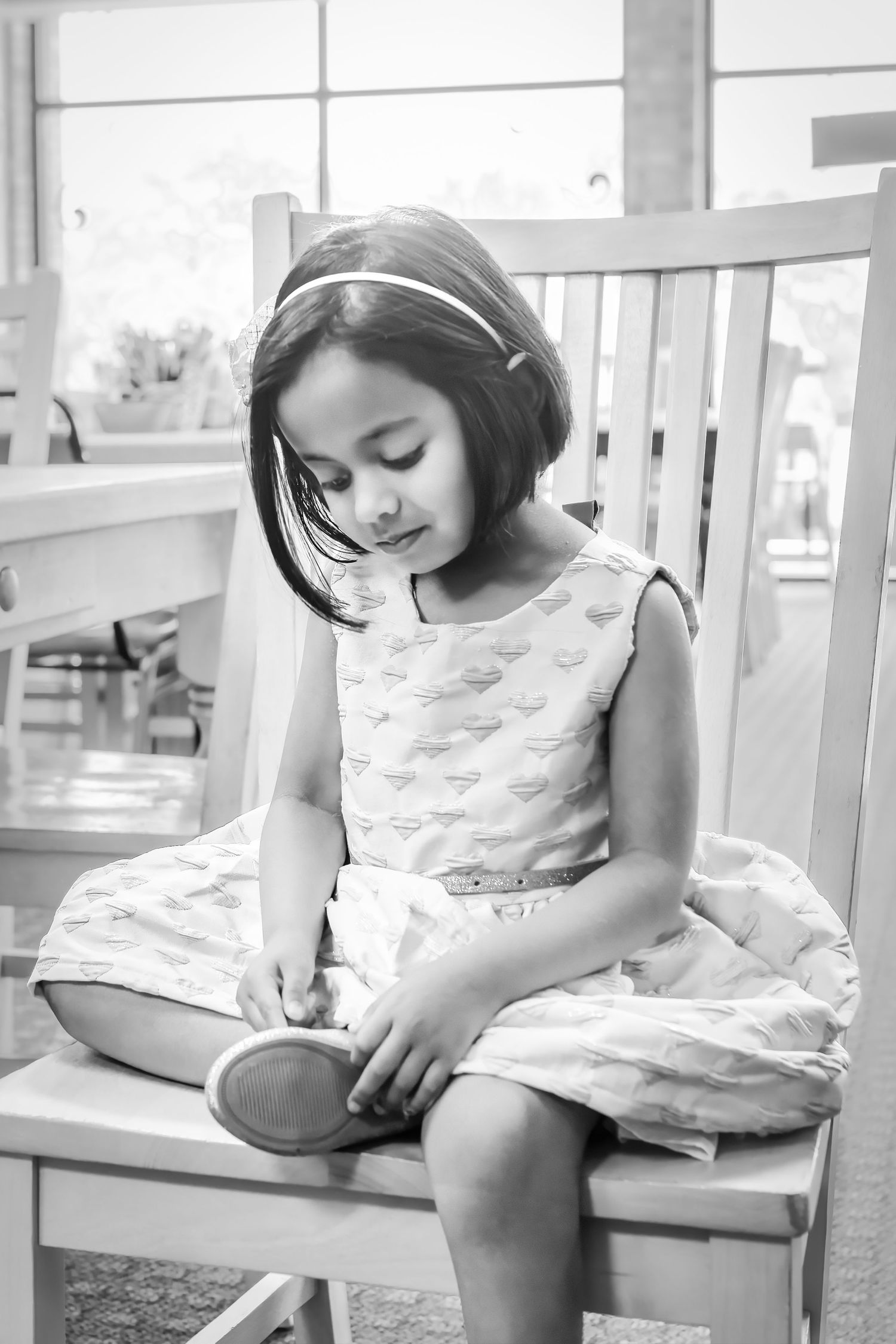 Black and white photo of a young girl in a patterned dress sitting on a wooden chair, looking down at an object in her hands.