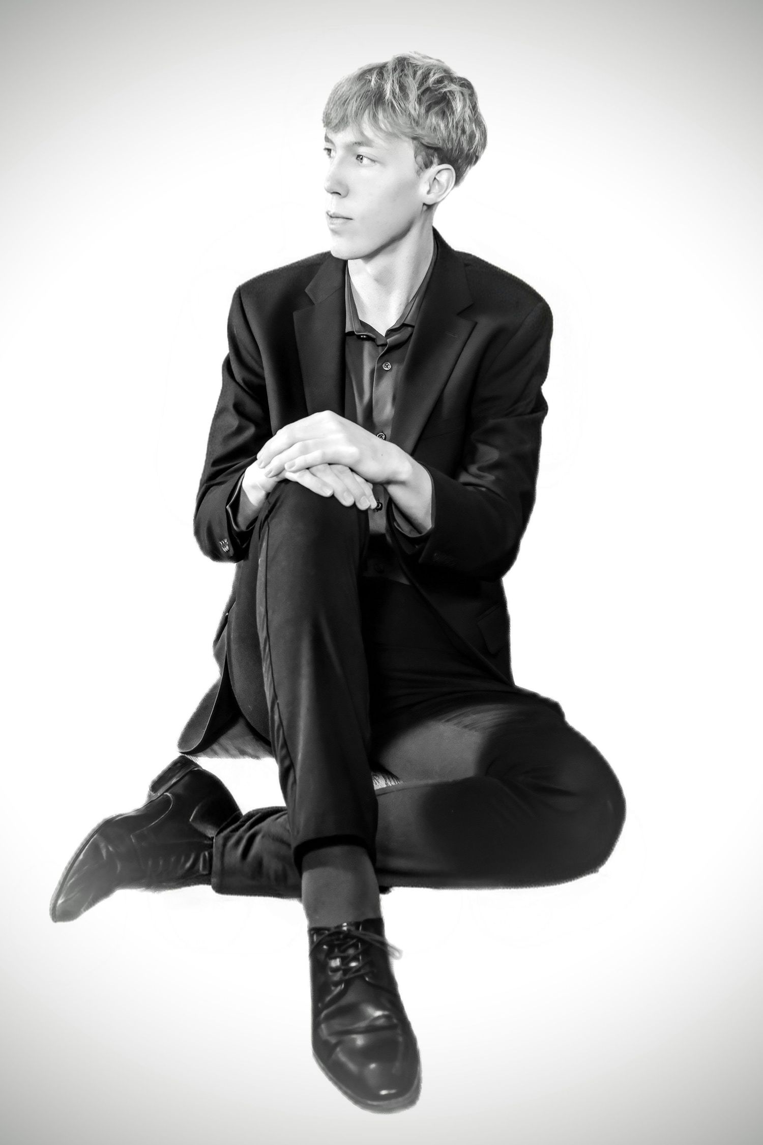 Black and white portrait of a young man in a black suit sitting cross-legged on a white background.