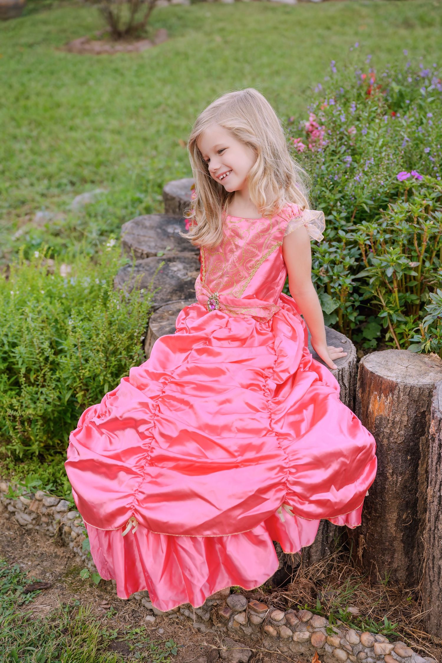 Little blonde girl in pink ball gown sitting on a wooden stump in a garden.