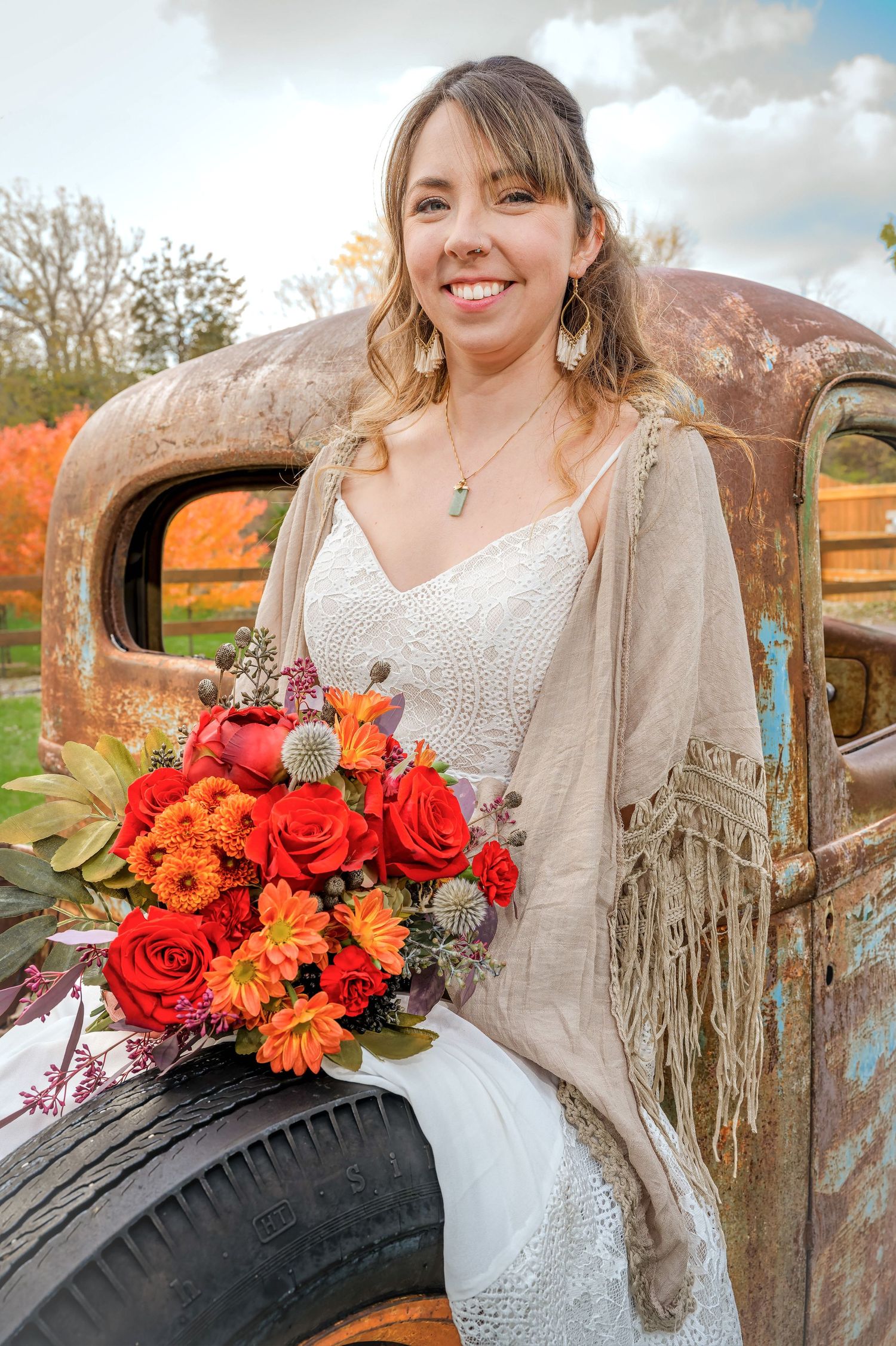 Smiling bride in white dress holding vibrant orange and red bouquet, seated on vintage rusty truck in autumn setting.