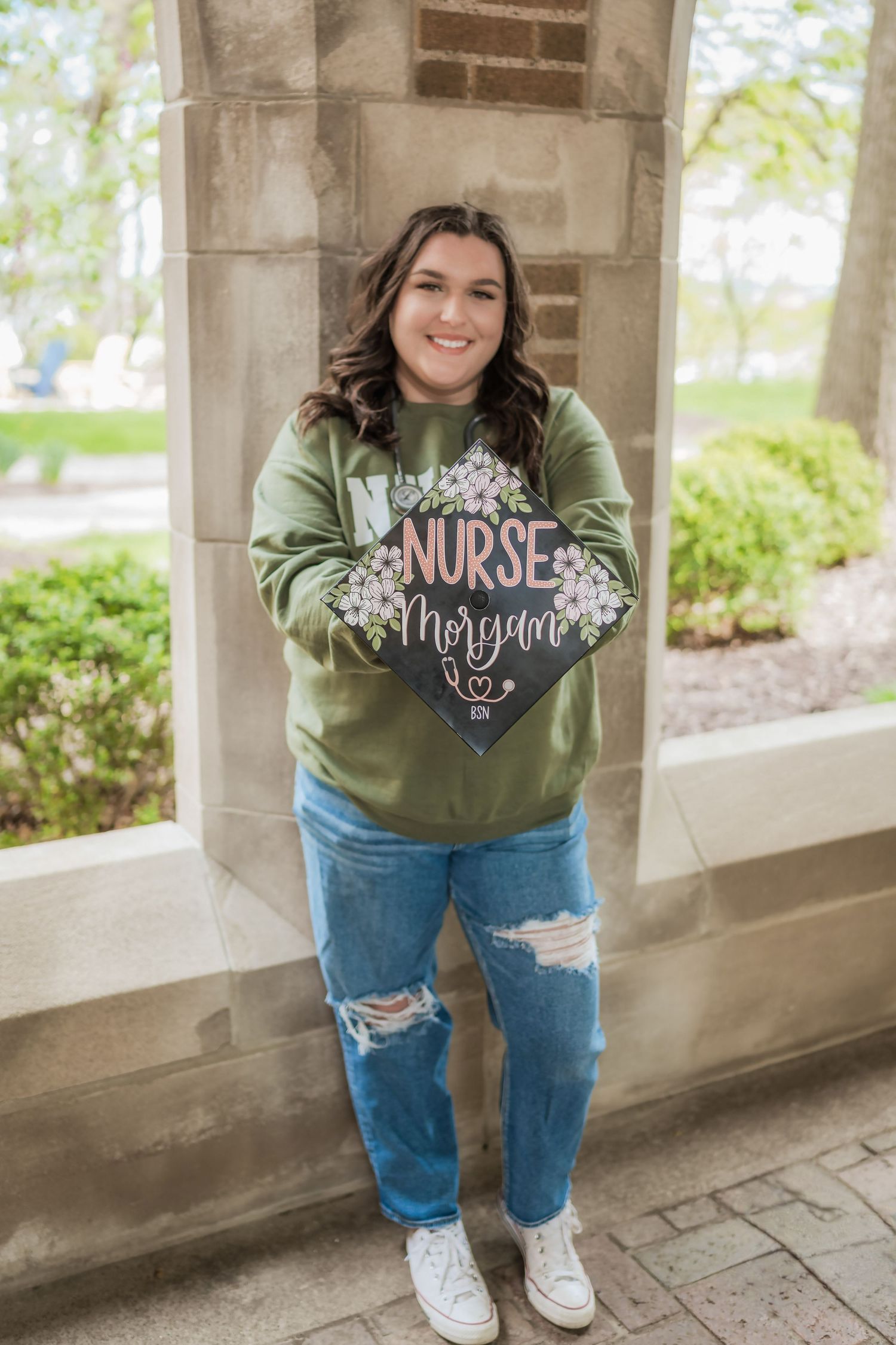 Smiling nursing graduate holding decorated mortarboard cap with name Morgan outdoors.
