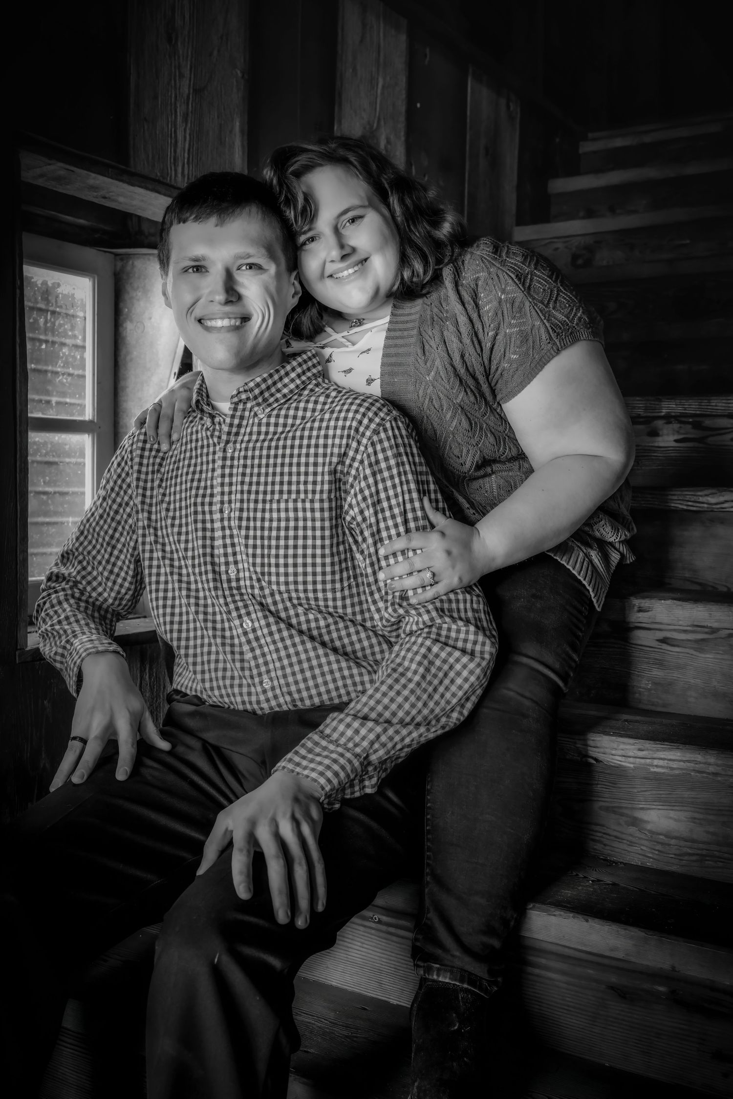 Black and white portrait of a smiling couple sitting on wooden steps inside a rustic log cabin.