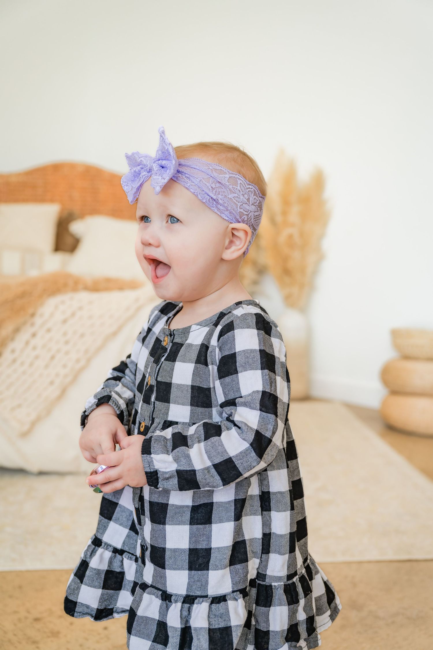 Adorable baby girl in plaid dress with purple bow headband laughing indoors.