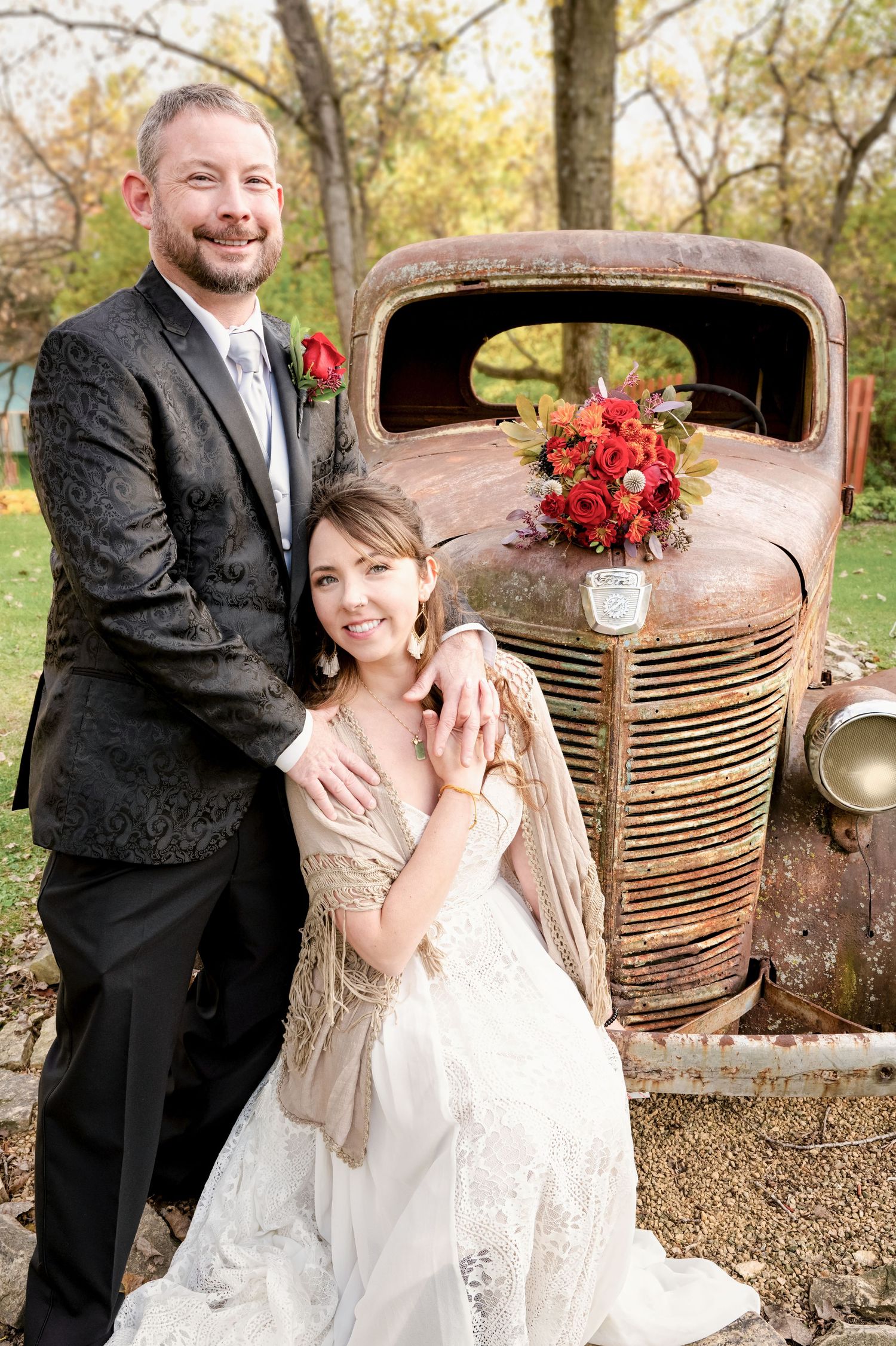 Wedding couple posing beside a rustic vintage truck with red floral bouquet, bride in lace gown.
