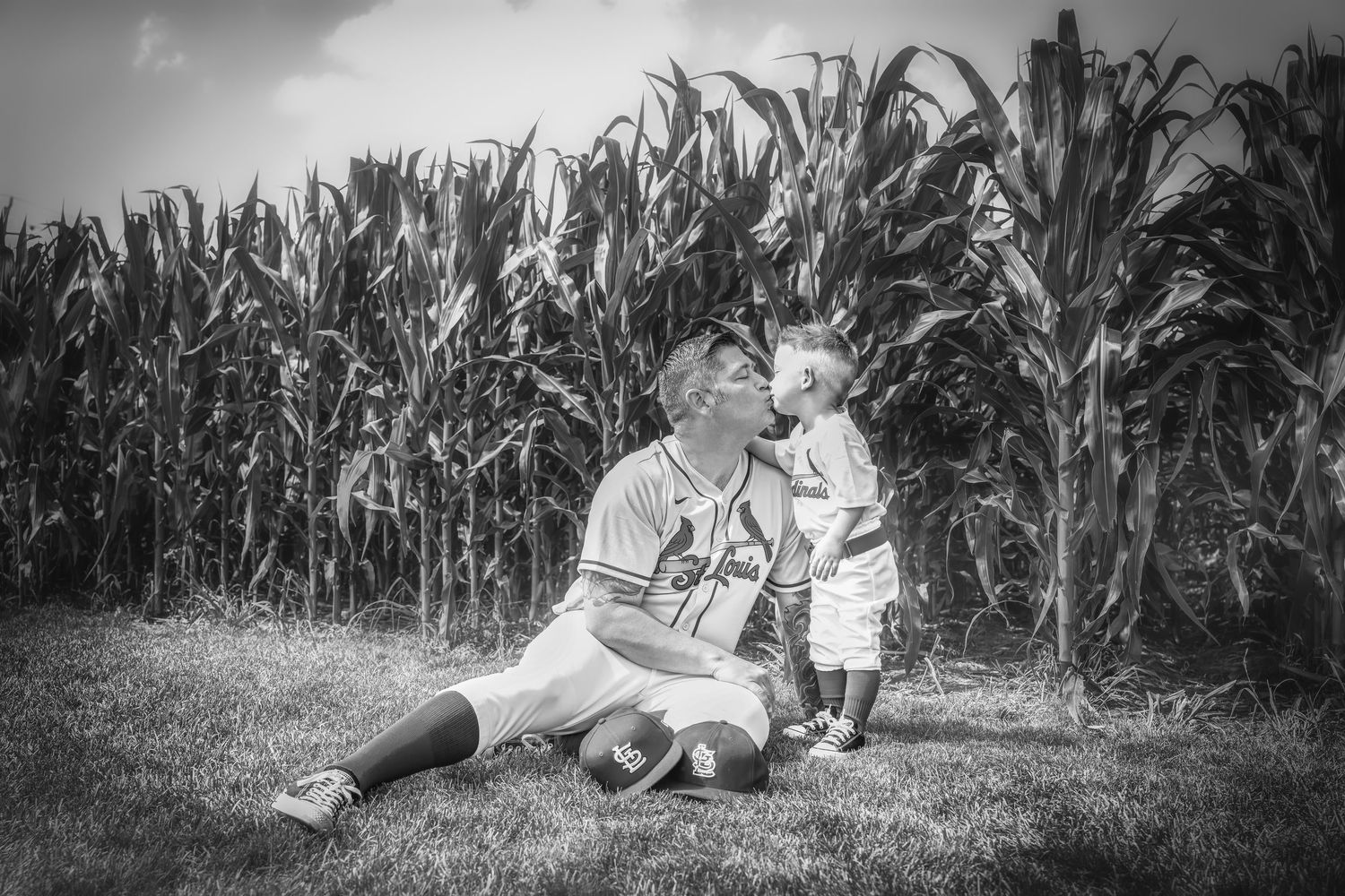 Black and white photo of a man in baseball uniform sitting with a toddler near a cornfield.