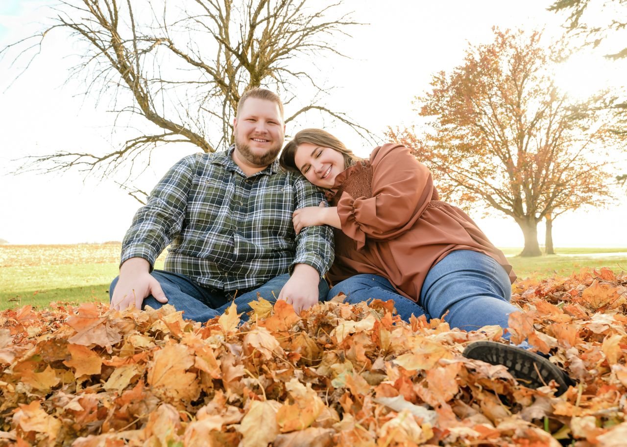 Couple sitting in autumn leaves outdoors, smiling, surrounded by fall foliage and trees in a sunny park.