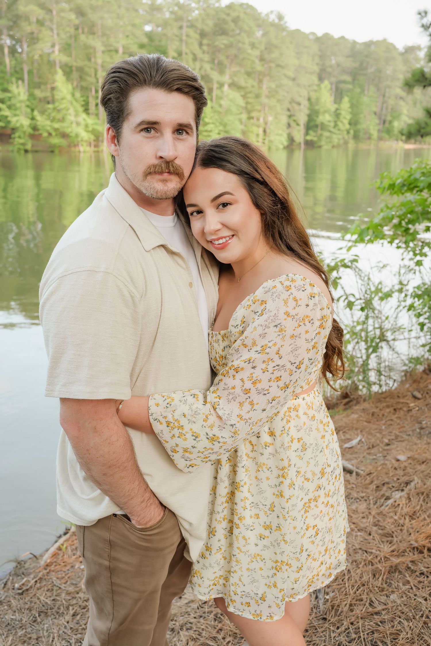 Couple embracing by a serene lake surrounded by lush green trees, woman in floral dress smiling at camera.