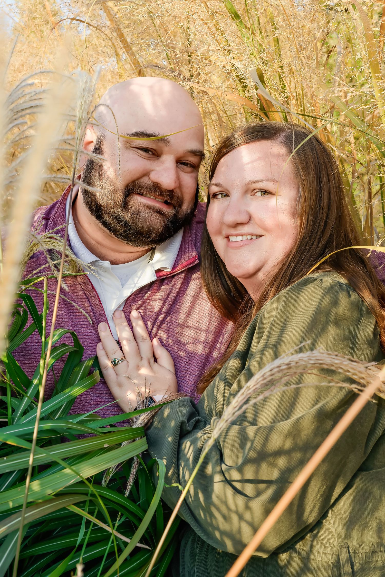 A smiling couple embraces among tall golden grasses in a warm outdoor engagement photo session.