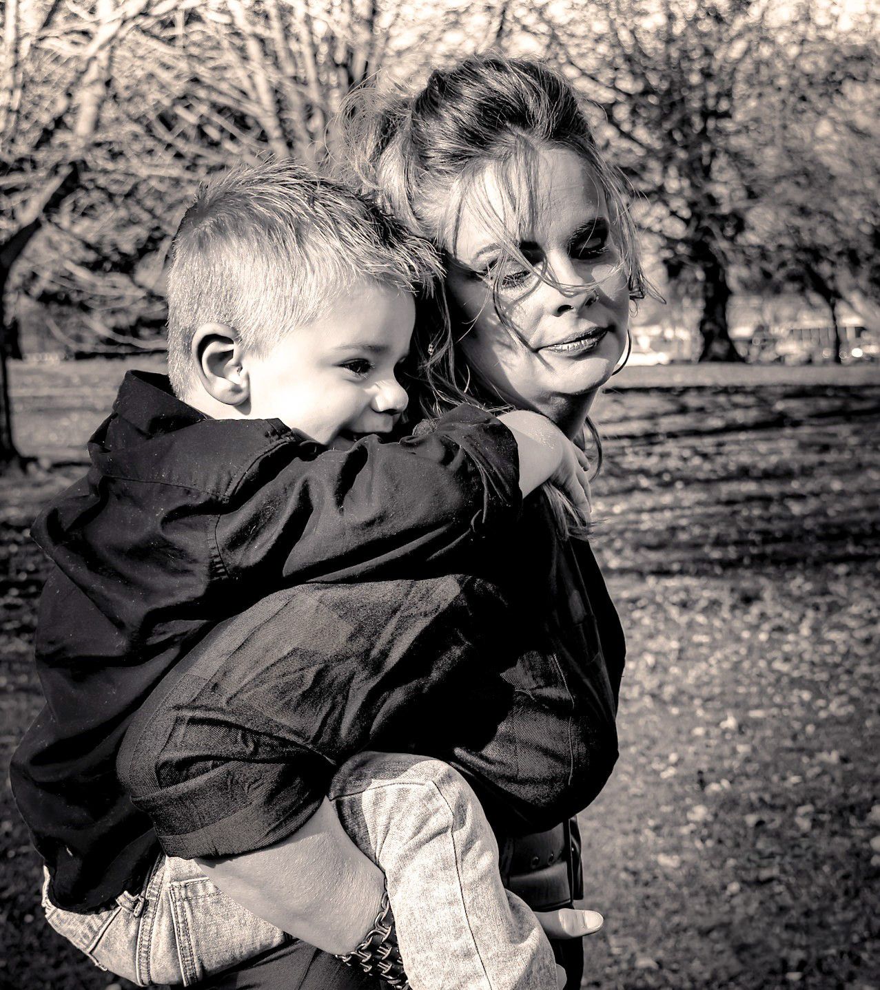 Black and white portrait of a smiling woman holding a young boy in an autumn park setting.