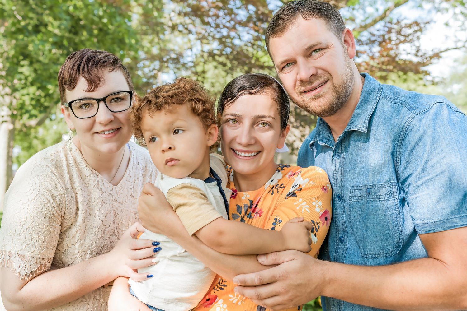 Happy family of four with young child posing together outdoors under green trees.