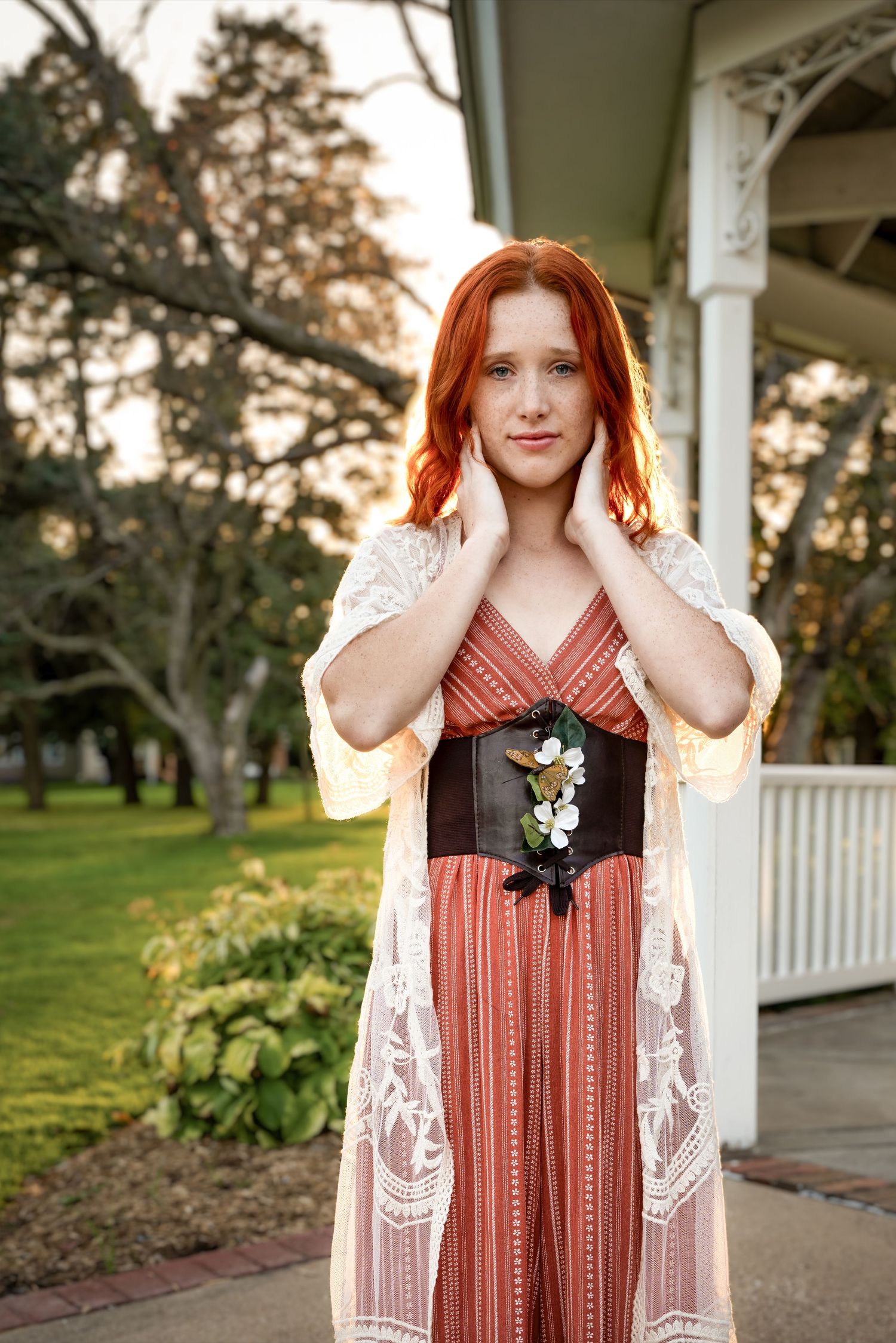 Elegant redhead woman in rust maxi dress and lace kimono posing on porch at sunset.