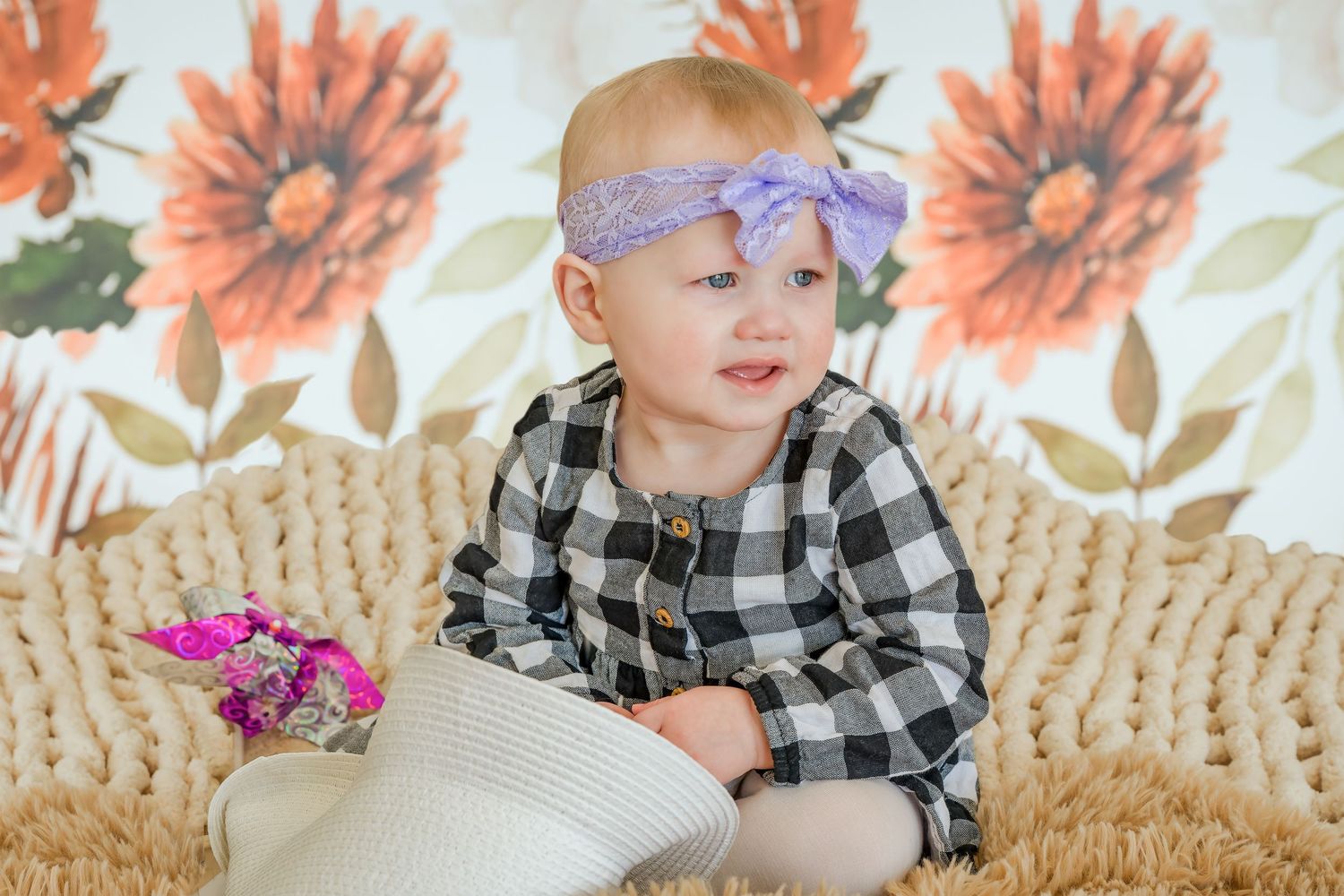 Cute baby in black checkered outfit wearing purple bow headband sitting on woven blanket.