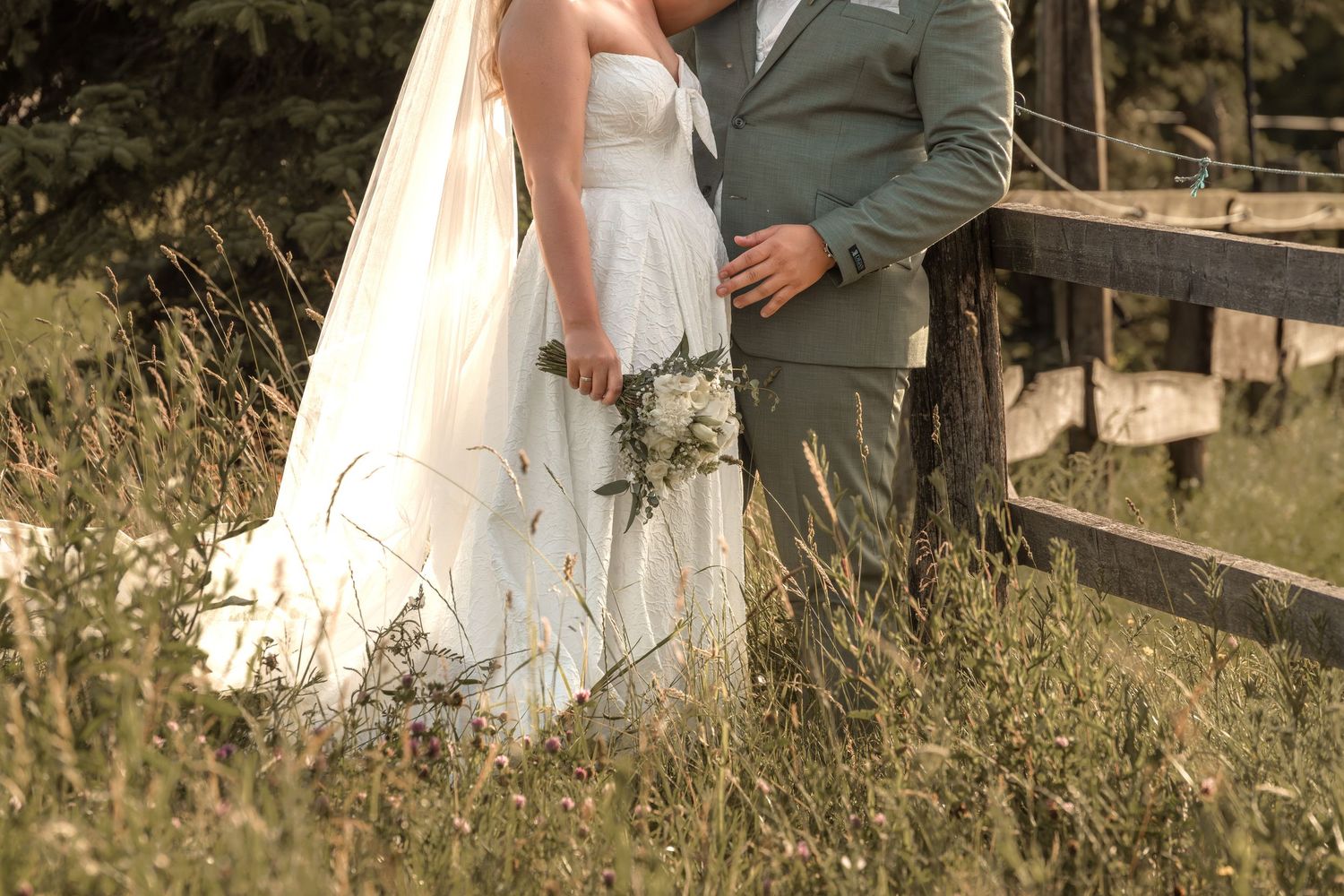 A bride and groom stand in a meadow, the bride holding a wildflower bouquet in a white gown beside a rustic fence.
