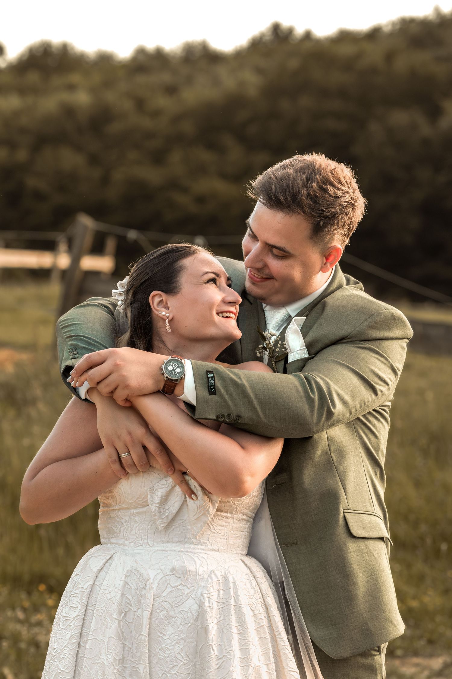A groom in a green suit embraces his smiling bride in a white dress outdoors during golden hour.