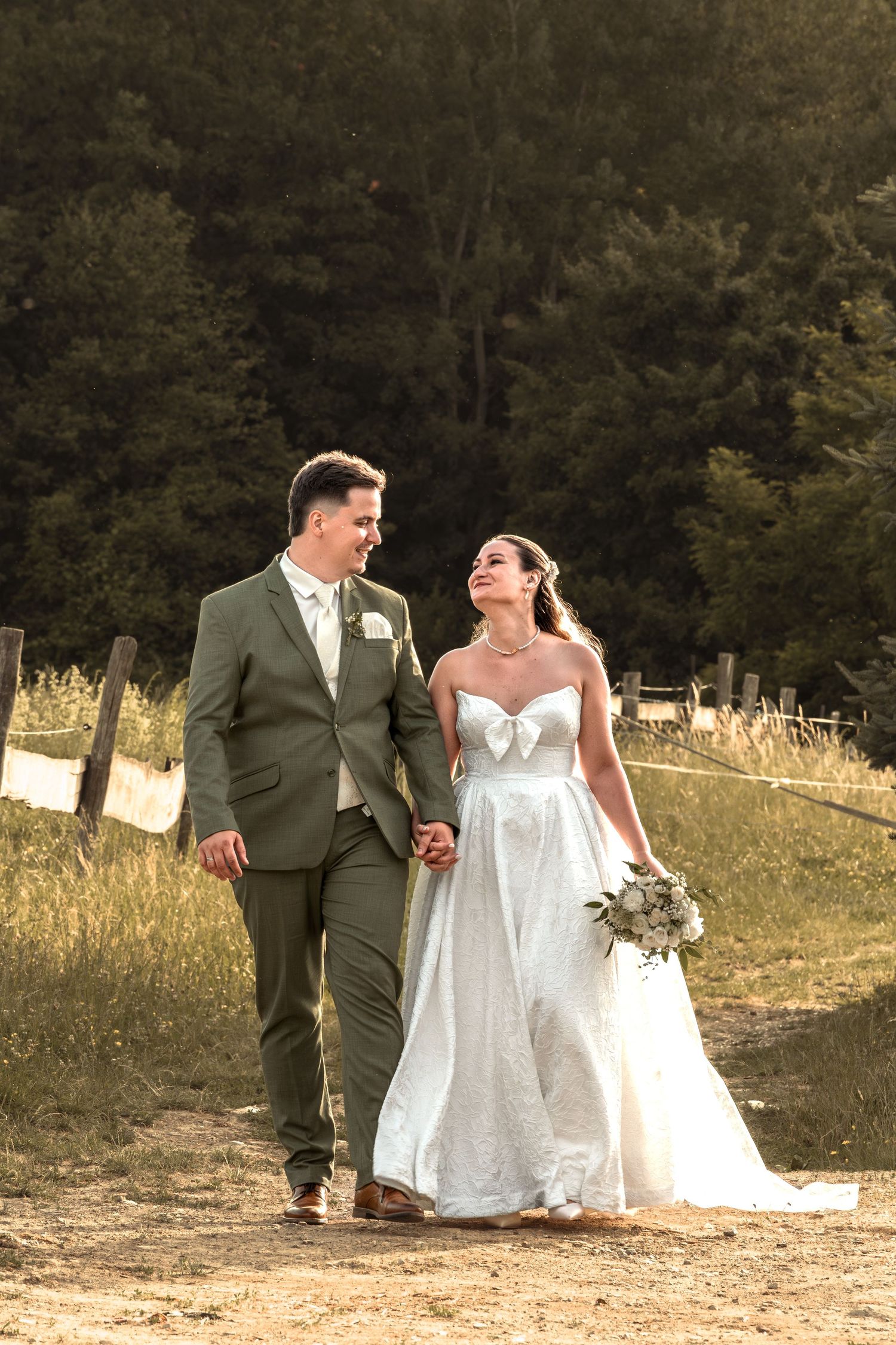 Bride and groom walking hand in hand through a sunlit field, smiling at each other in elegant wedding attire.