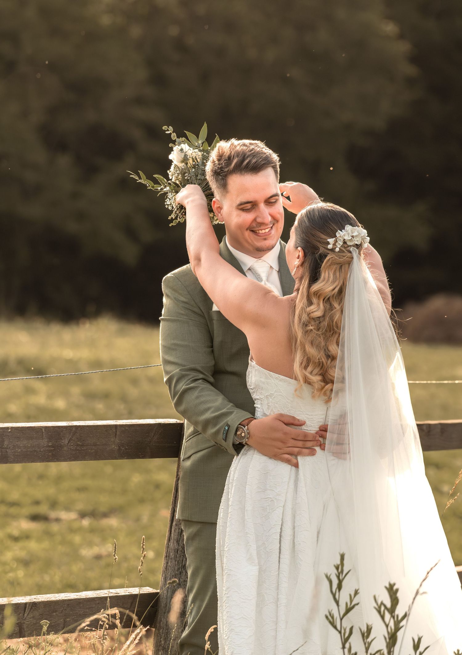 Bride and groom embracing outdoors at golden hour, bride holding bouquet aloft, groom in sage suit, pastoral fence backdrop.