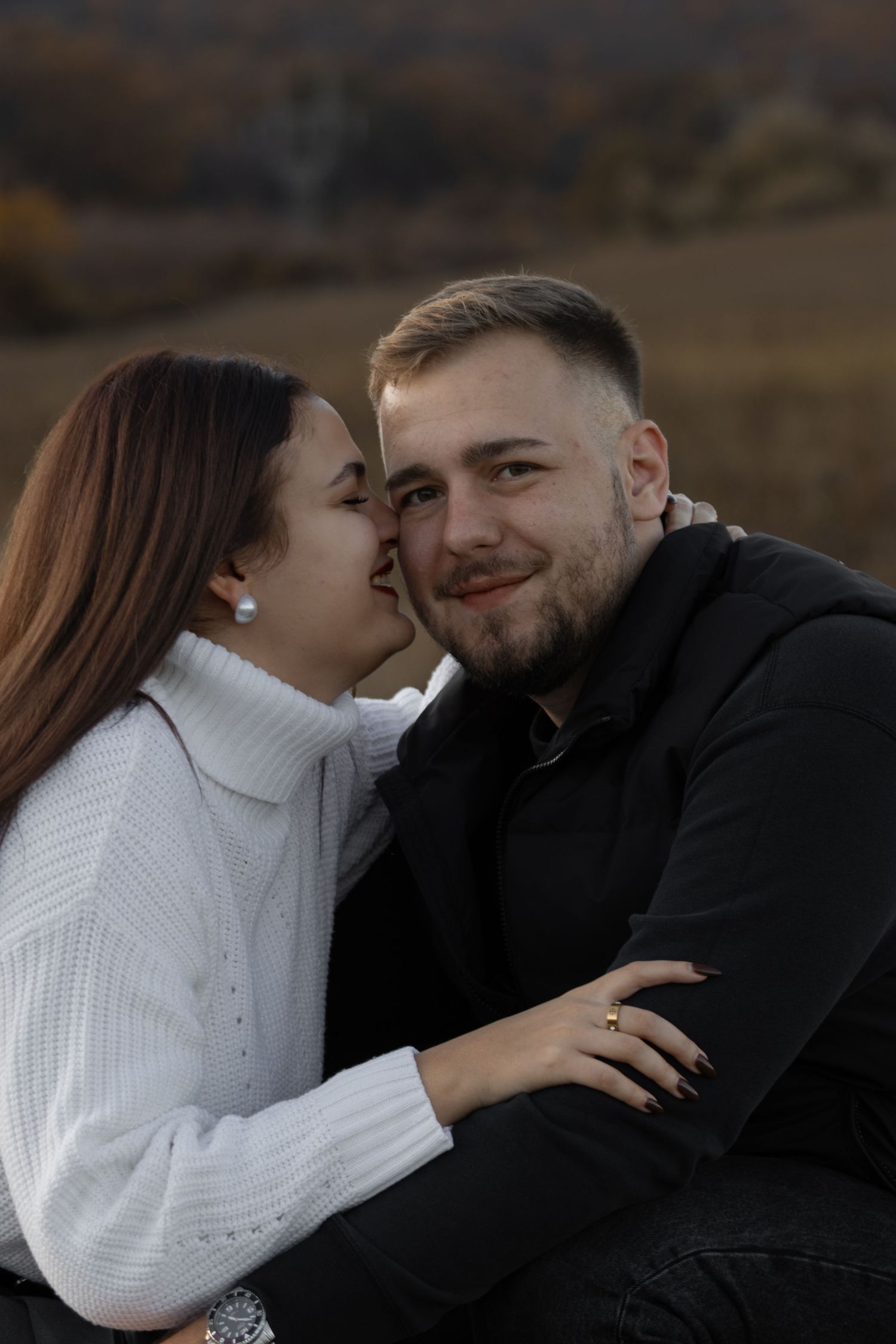 nő fehér pulóverben odasúg valamit a mosolygó férfinak egy őszi tájon