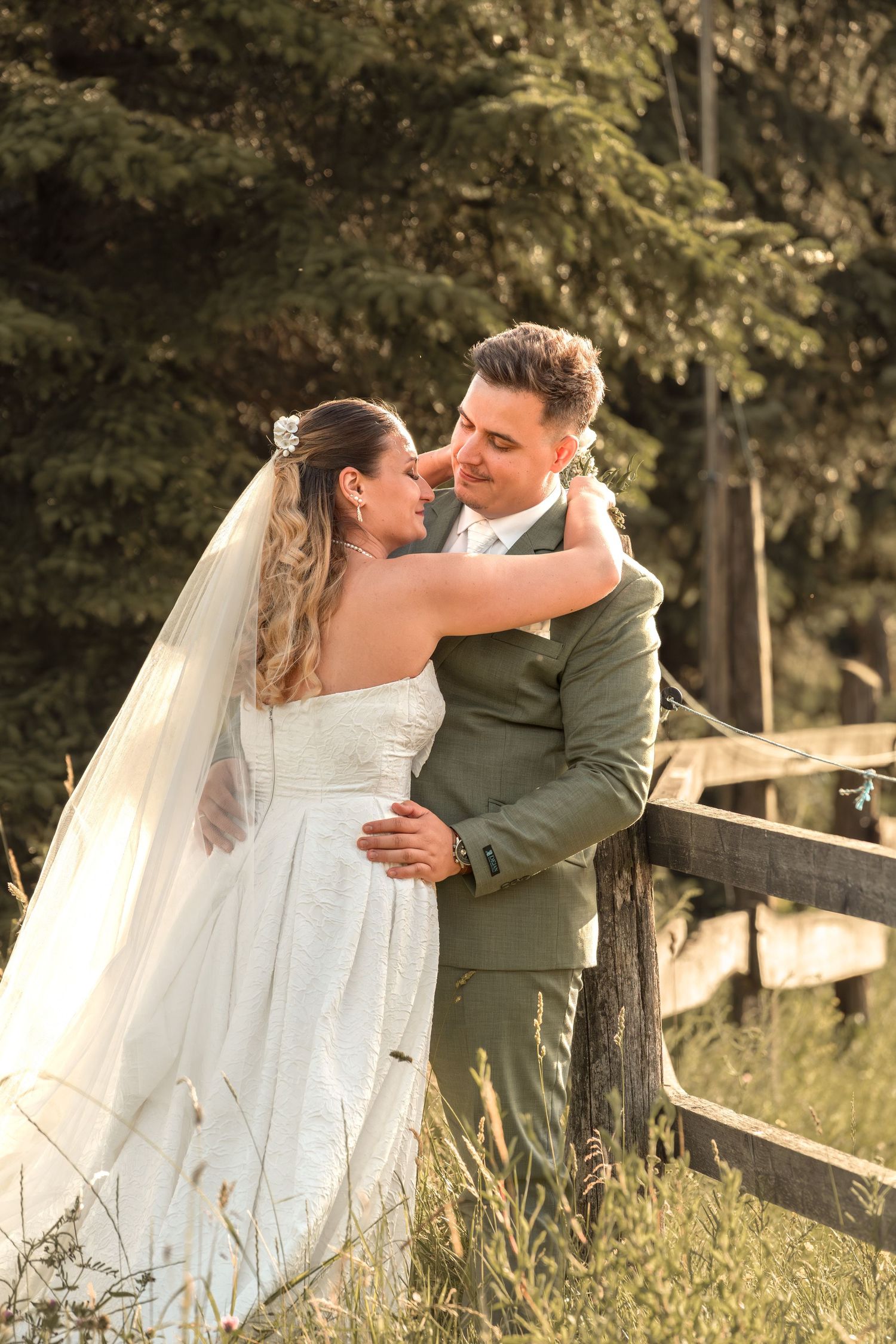 Bride in white gown and veil embraces groom in olive suit near rustic fence surrounded by lush green trees.