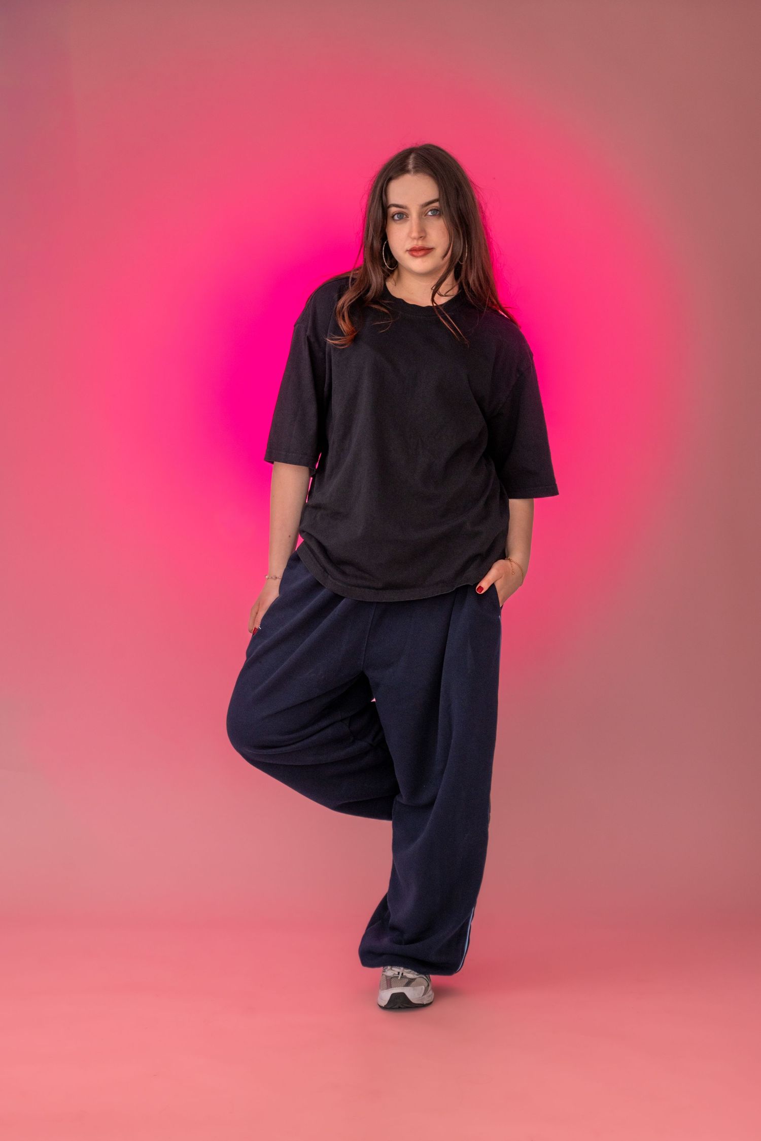 Egy nő fekete oversized pólóban és sötétkék bő szárú nadrágban magabiztosan pózol rózsaszín neon háttér előtt.