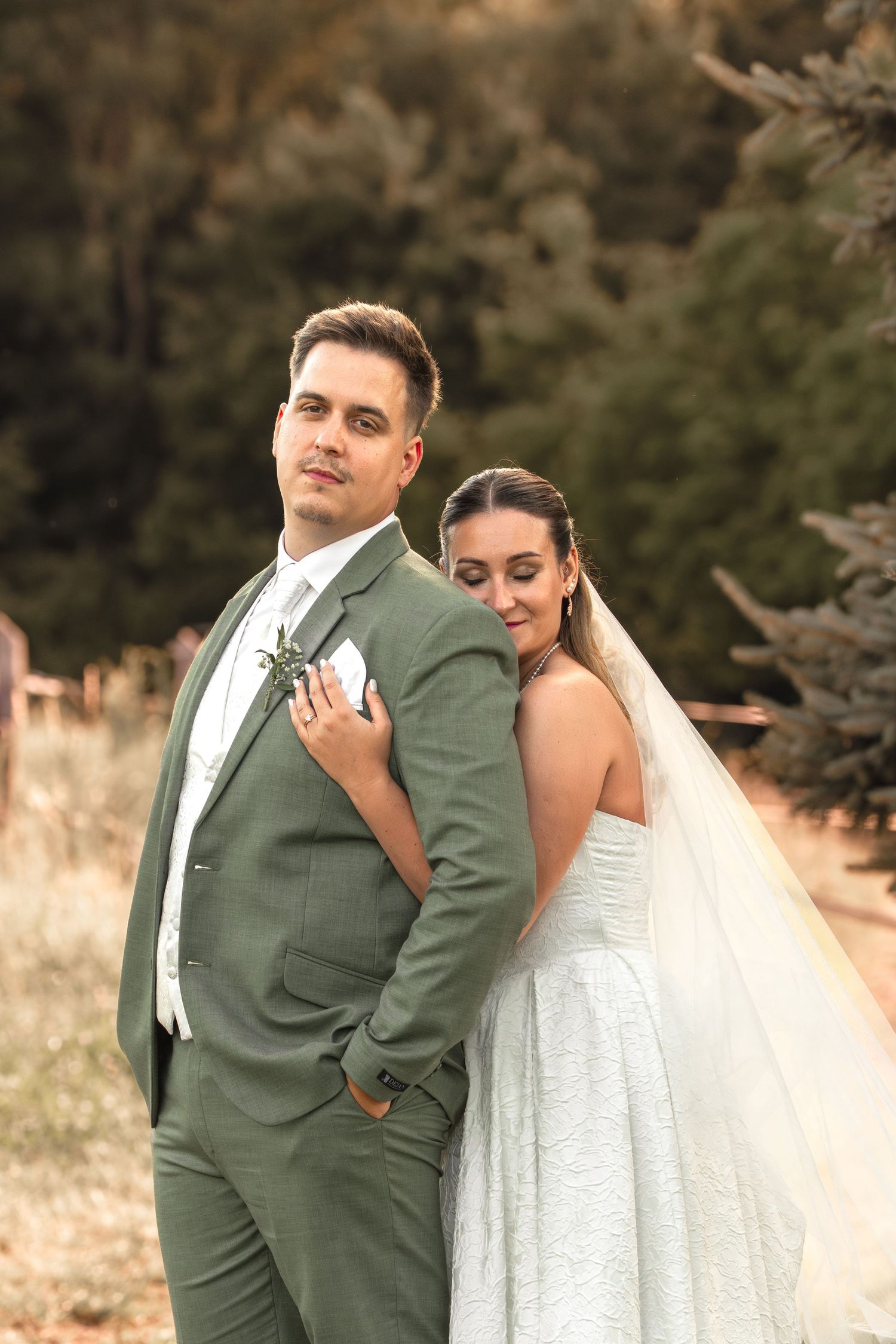 Bride in white gown and veil leans on groom in sage green suit, posing outdoors with trees in background.