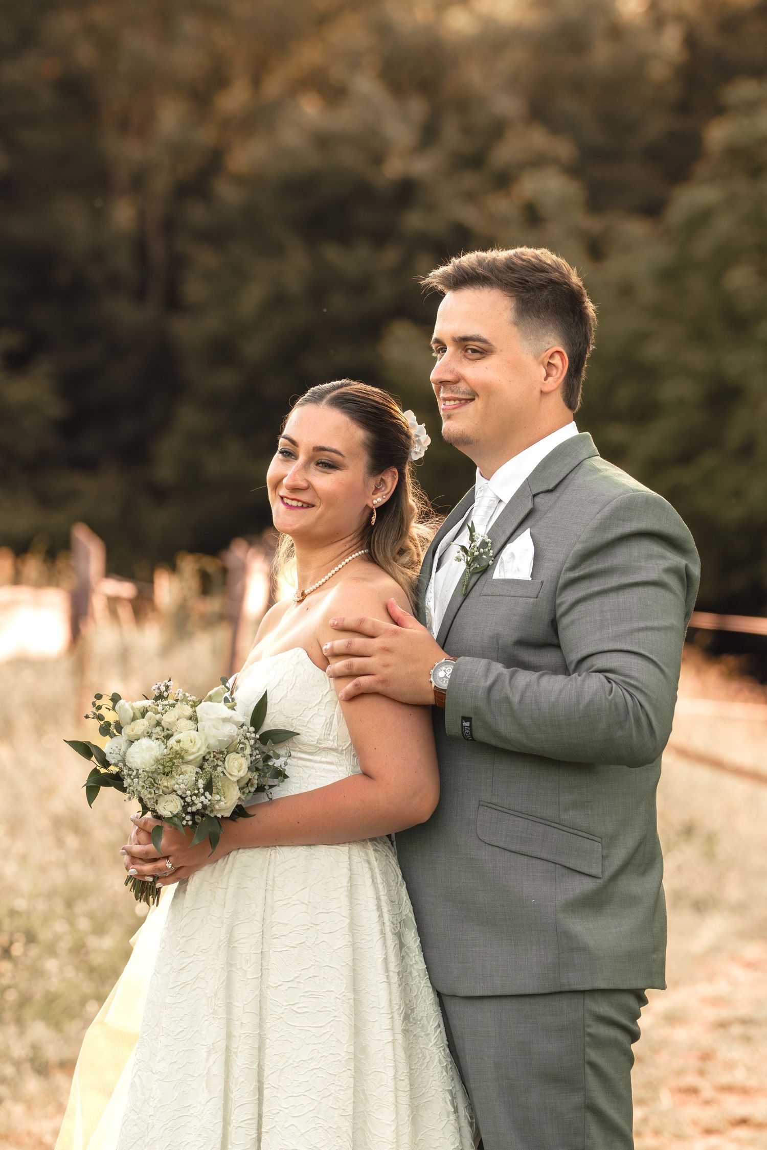 Smiling bride holding white bouquet with groom in gray suit embracing her outdoors at golden hour.