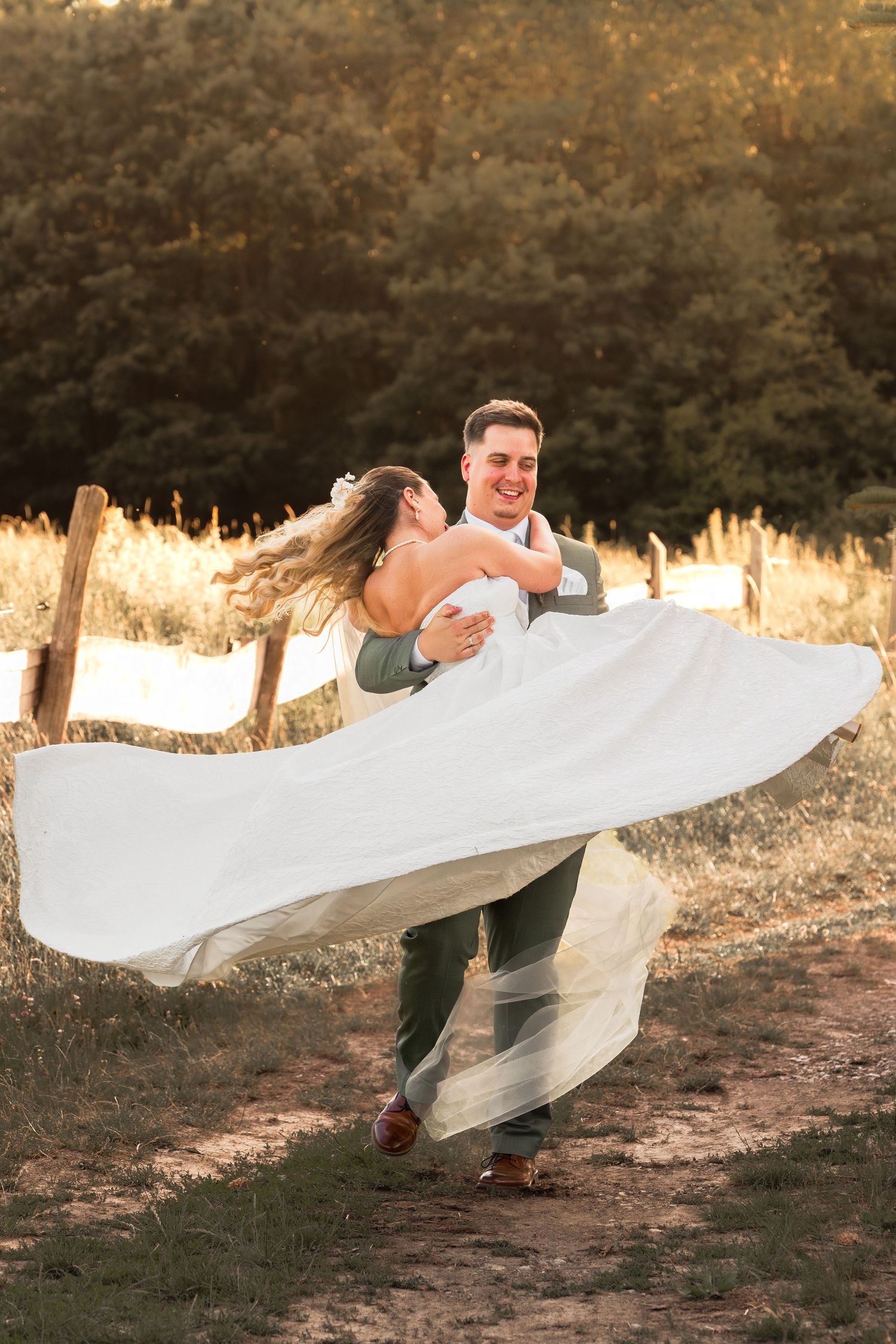 Groom spinning bride in white flowing dress outdoors at golden hour near rustic fence.