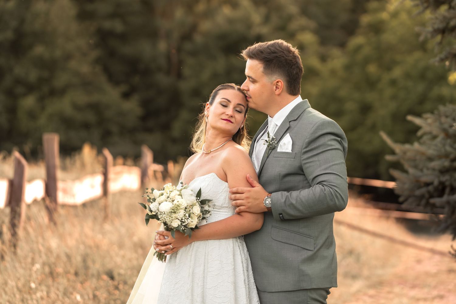 Bride and groom embracing outdoors, she holds white bouquet in wedding dress, he wears gray suit, forest backdrop.
