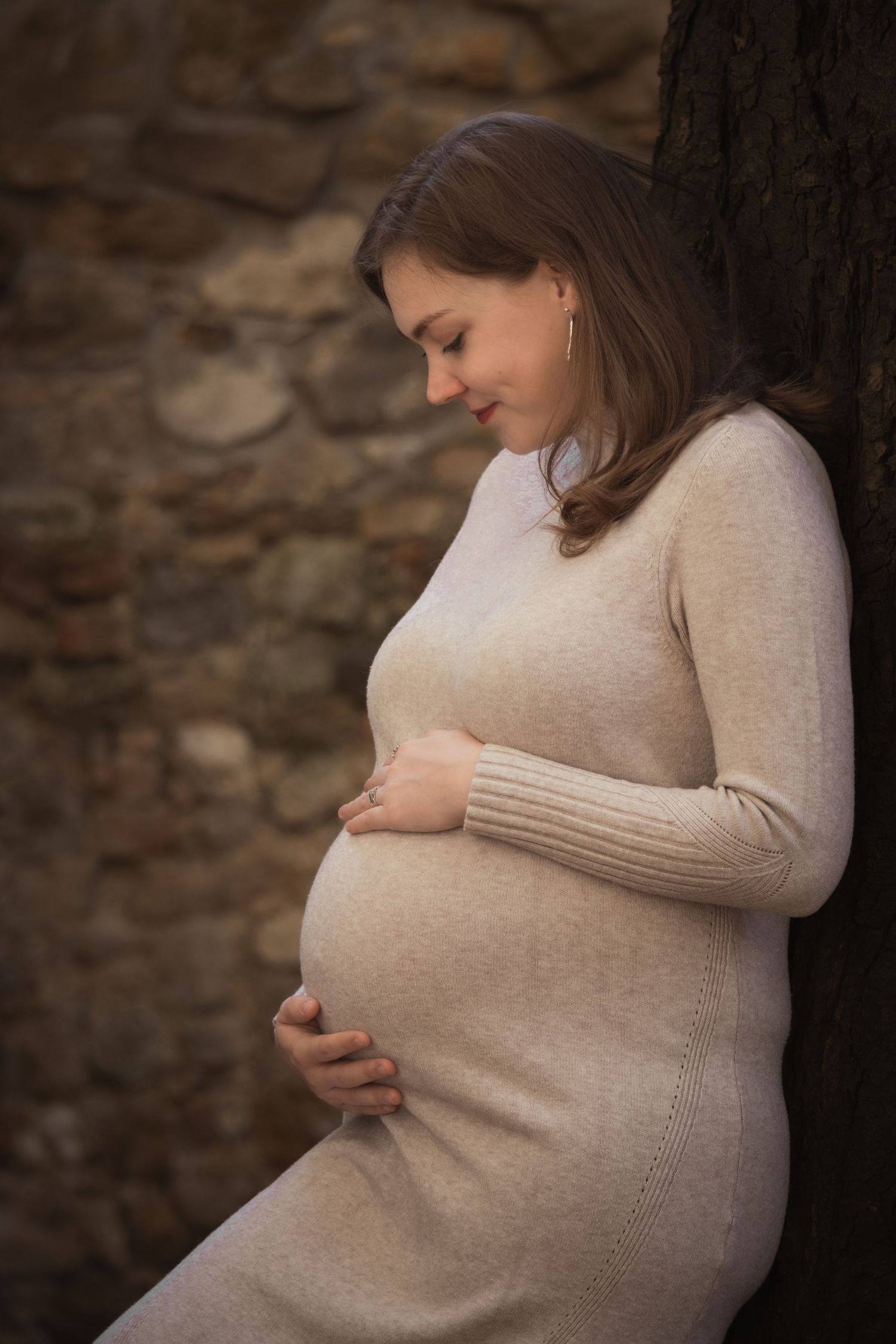 kismama bézs ruhában fának támaszkodva, pocakját ölelve