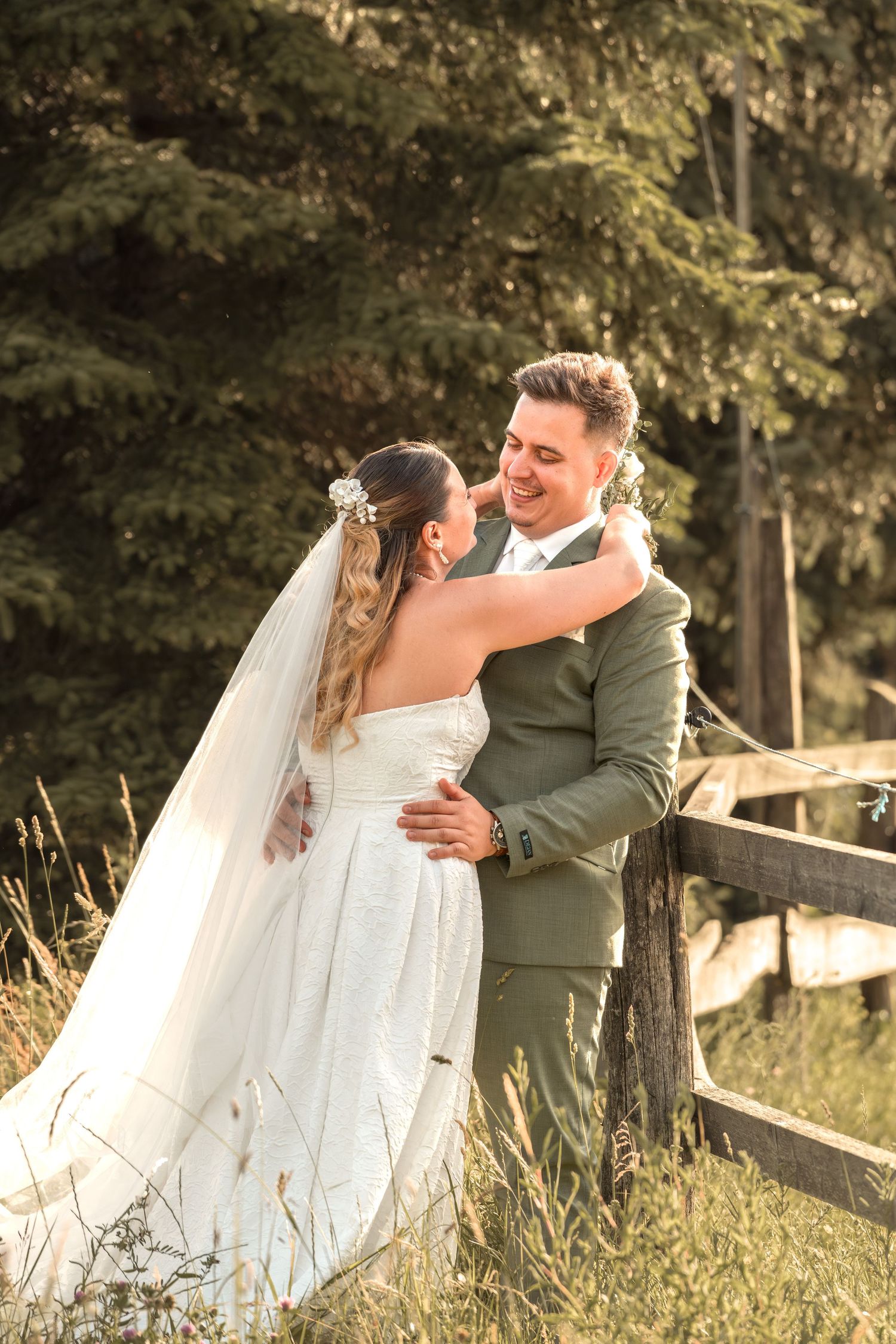 Bride and groom embracing outdoors near a wooden fence, surrounded by lush green trees on their wedding day.
