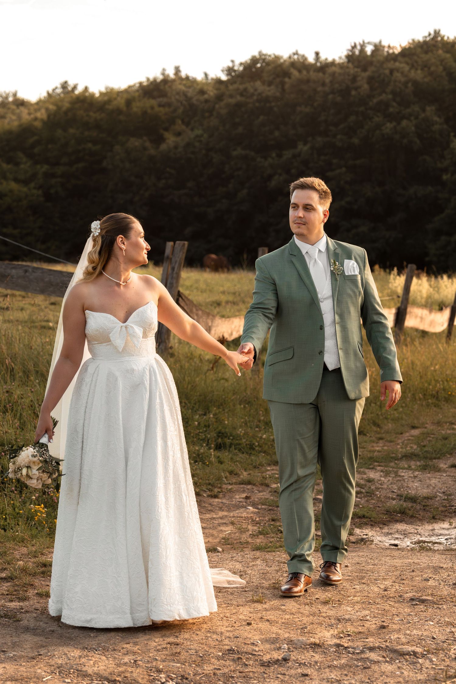 Bride in white gown and groom in green suit hold hands walking outdoors at golden hour.
