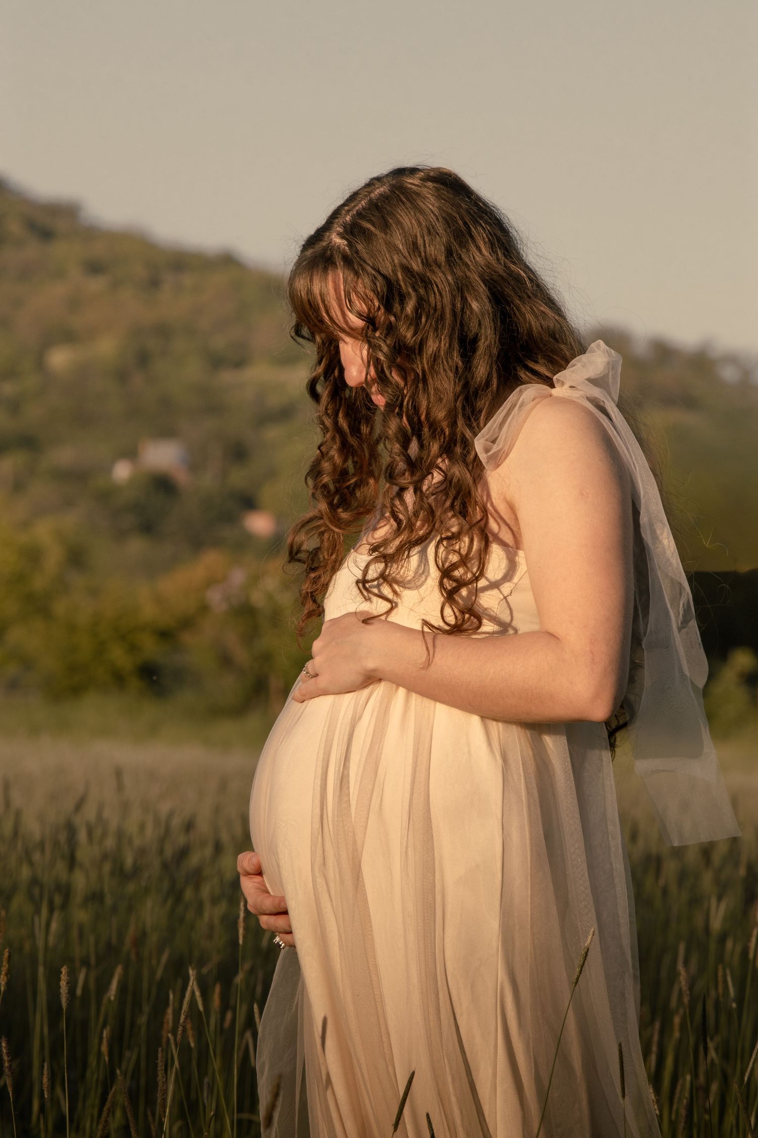 kismama áttetsző krémszínű ruhában, kezét a pocakján tartva naplementében, dombos tájjal a háttérben