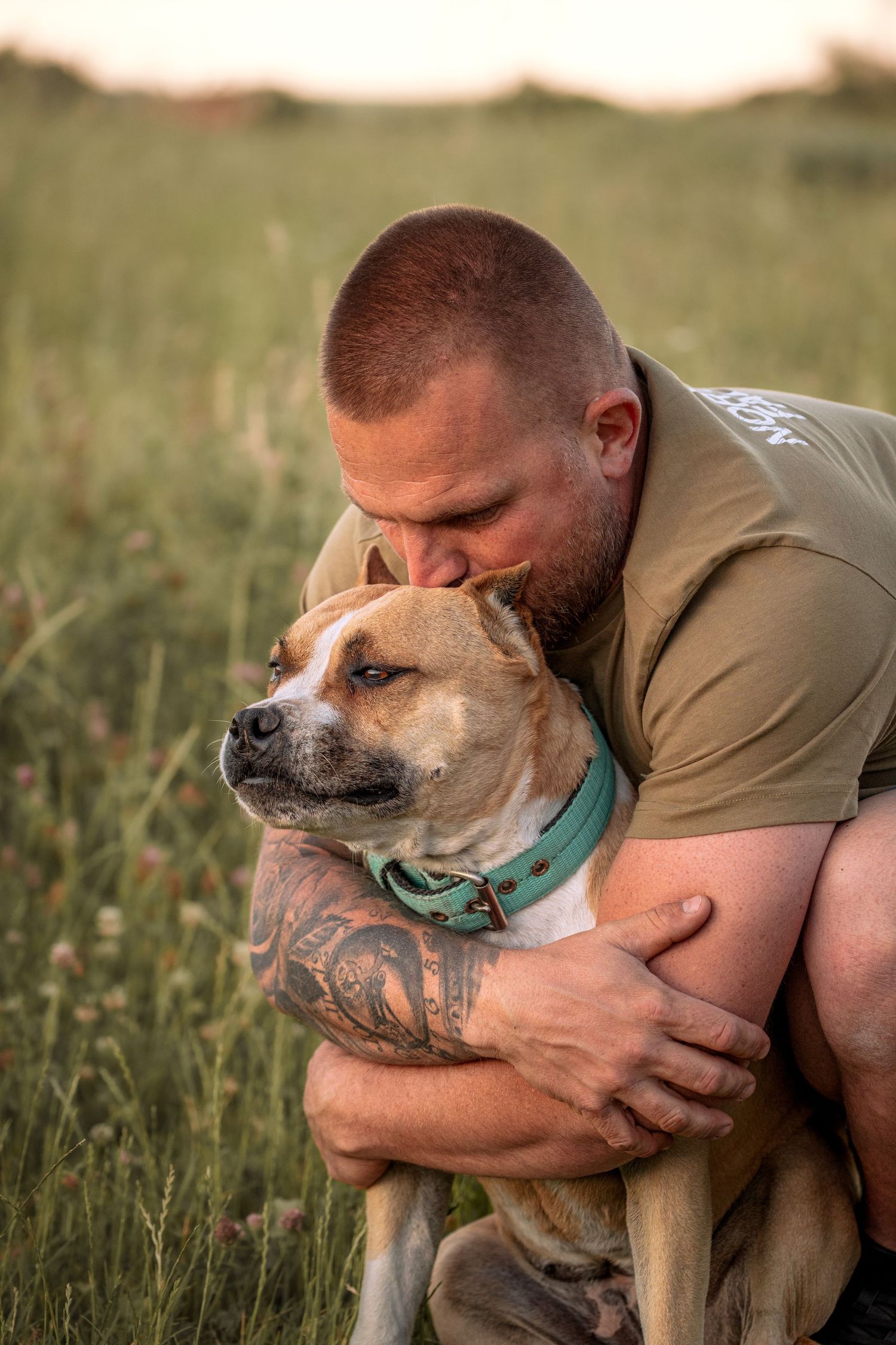 Tattooed man hugging and kissing his tan pit bull dog in a green field at sunset.