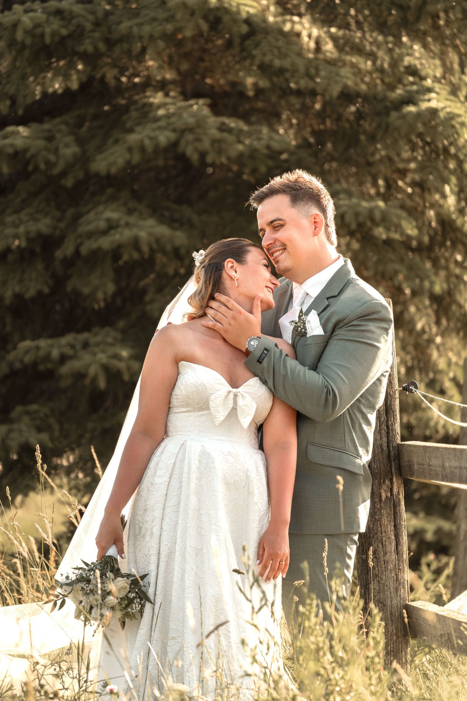 A smiling groom in a gray suit tenderly holds his bride's face as she laughs in a white strapless gown outdoors.