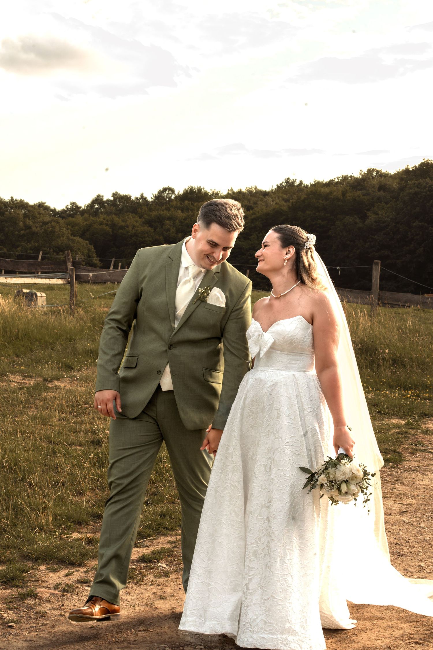 A bride and groom walk hand in hand through a countryside field, she in white lace, he in a sage green suit.